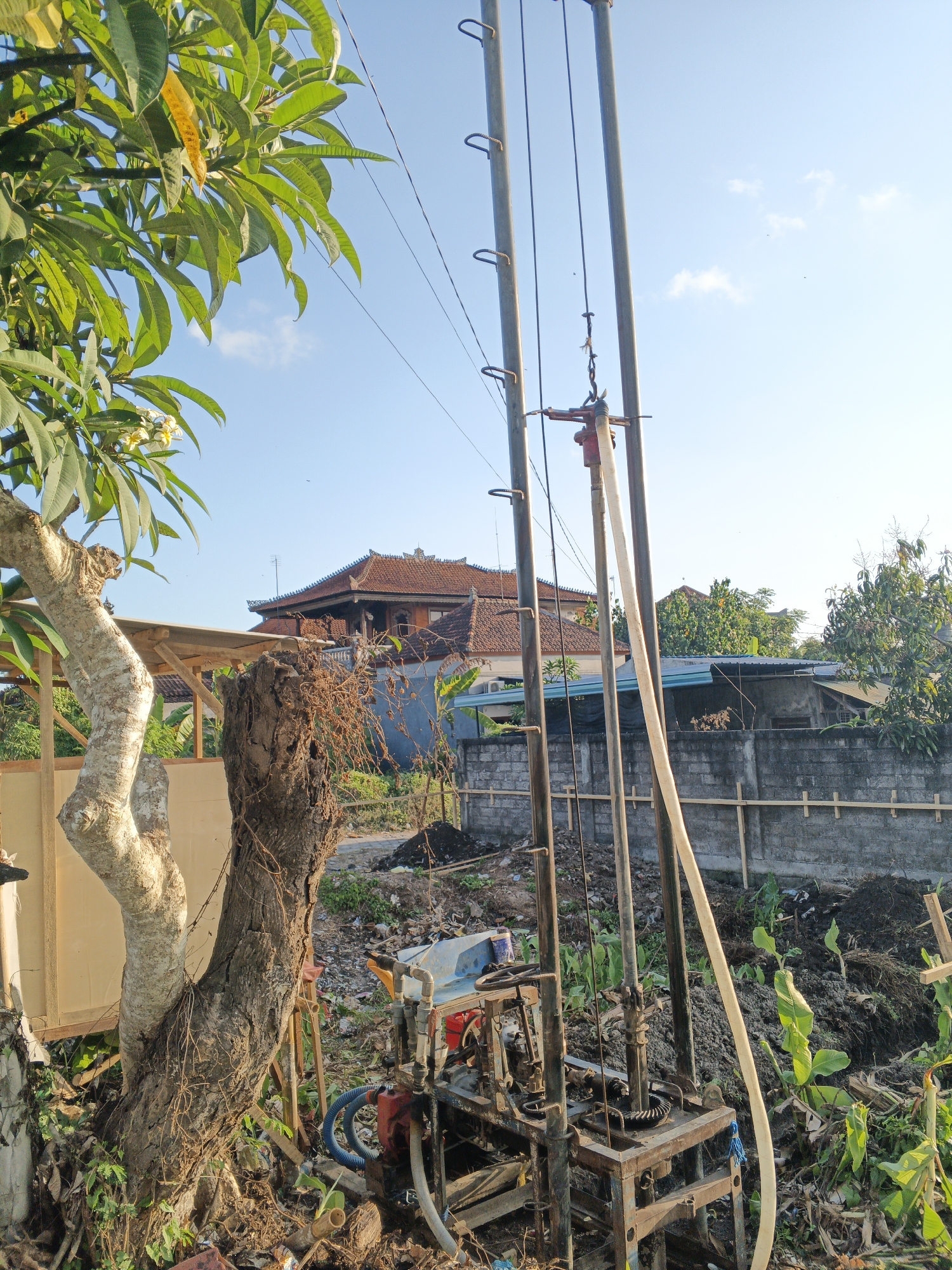 The image depicts a well-drilling operation in a semi-rural area of Sukawati, Indonesia. A metal drilling rig with several vertical poles stands prominently in the center, with a water hose attached and extending upwards. Below the rig, a red pump is visible, connected to blue tubing. The immediate surroundings are cluttered with debris, including cut tree branches, soil, and various discarded items. To the left, a large tree trunk with exposed roots and a smaller, lighter-barked tree are situated. In the background, traditional Indonesian houses with tiled roofs are visible, along with a grey concrete wall topped with wooden planks. Green foliage and vegetation are abundant throughout the scene. The sky is clear and blue, with a few scattered clouds. The lighting suggests it is daytime, likely late afternoon due to the warm tones. There are no people present in the image.