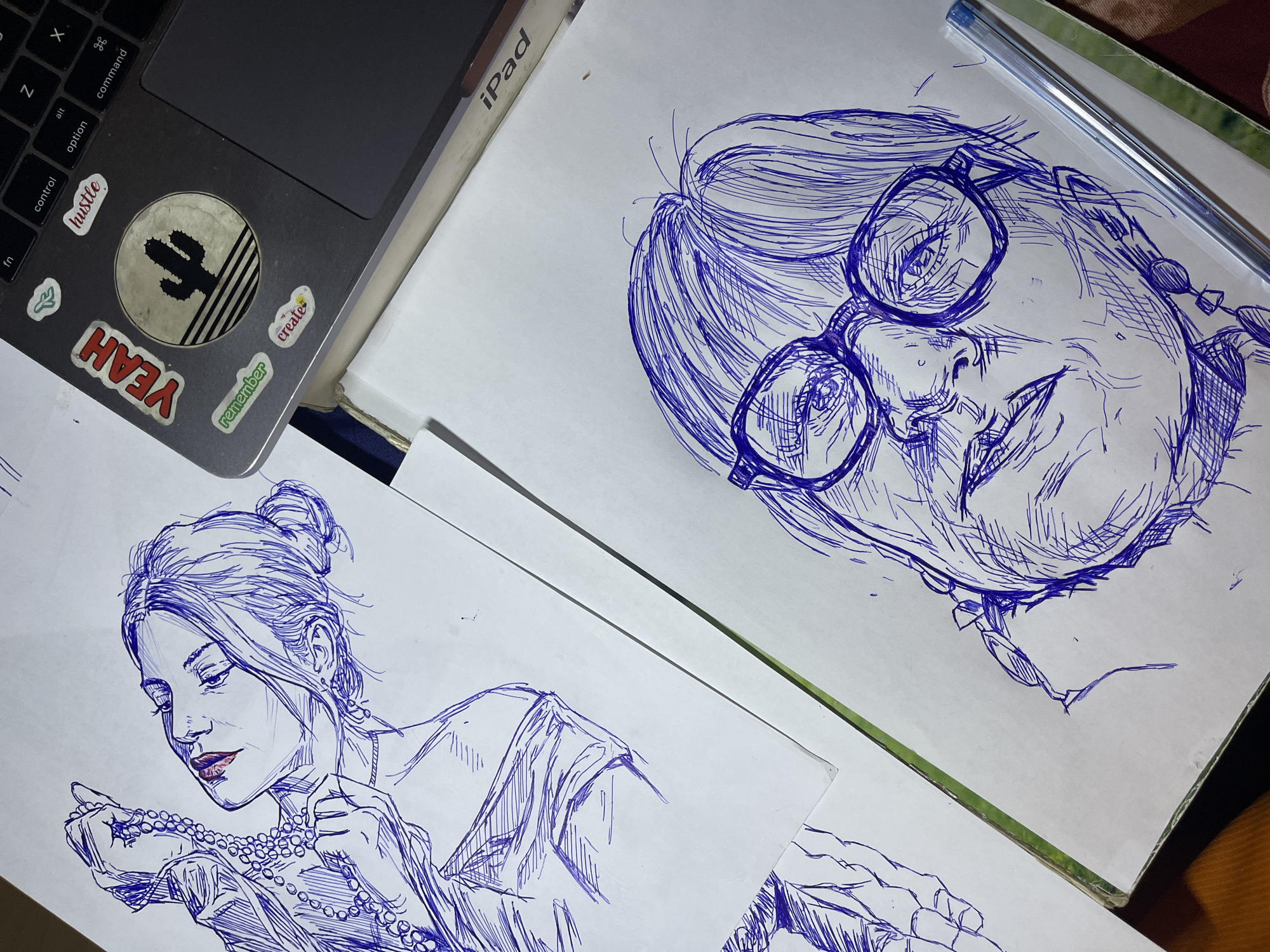 The image displays two blue ink sketches on white paper. The top sketch is a portrait of a woman holding a strand of pearls to her neck. She is looking upwards with her eyes closed and lips slightly parted. Her hair is pulled back in a bun. The bottom sketch is a portrait of a person wearing glasses, facing forward with a serious expression. The sketches are laid out on a surface that includes a laptop keyboard with stickers, and a portion of an iPad. The overall scene is illuminated by artificial light, creating a dimly lit environment. There is no specific information within the image to indicate a location in Jos, Nigeria.