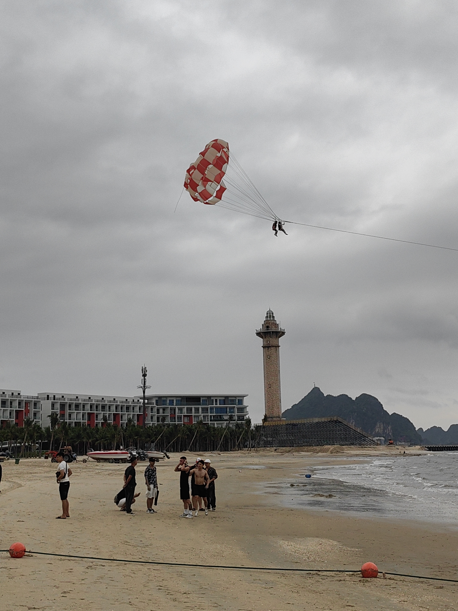 On a beach in Bai Chay Ward, Vietnam, a group of people stands near the shoreline. Two people are parasailing in the sky, connected to a boat by a long cable. The parasail is red and white checkered. Behind the group, there is a building and a lighthouse. The sky is cloudy. The beach is sandy and wet.