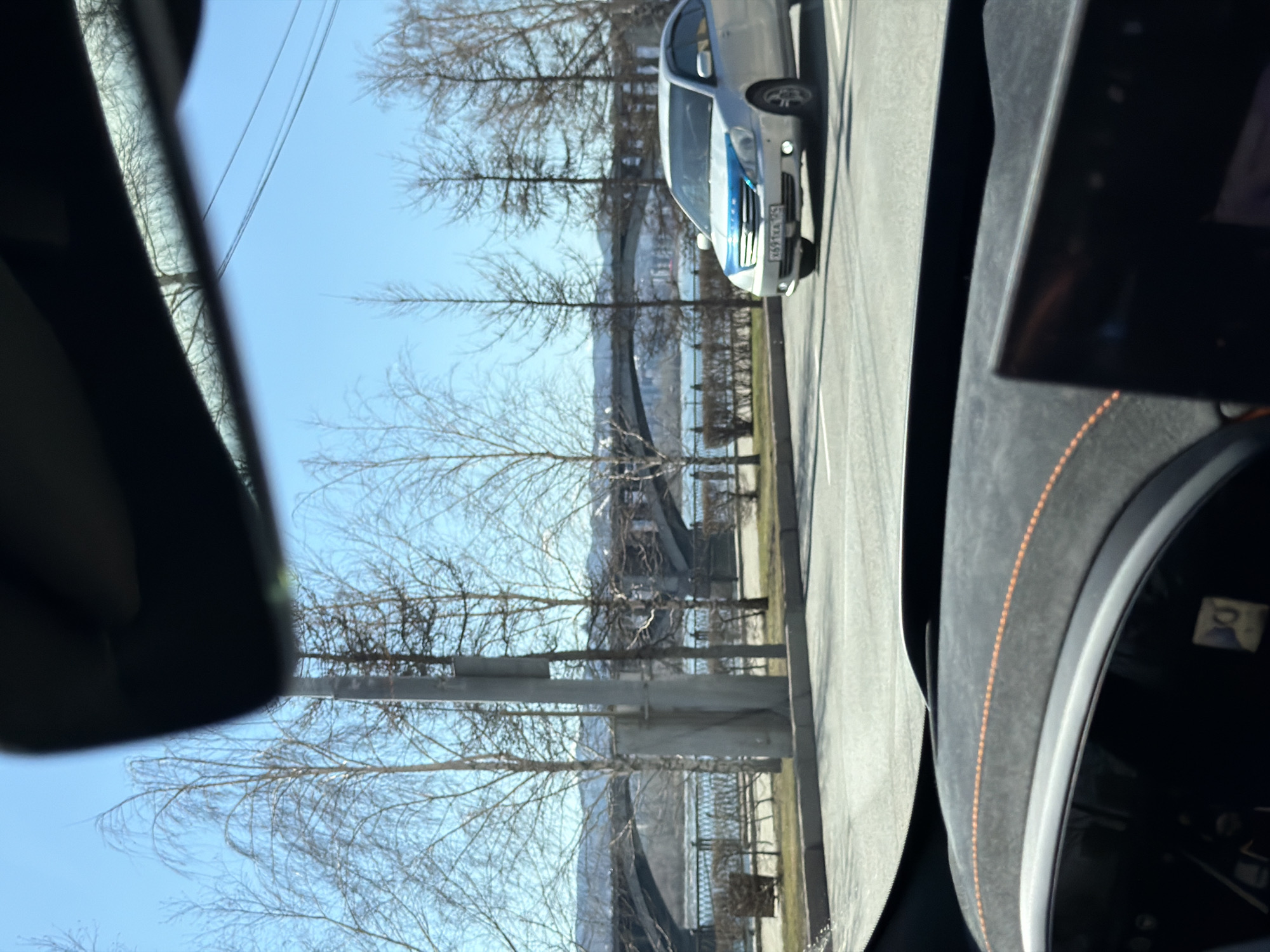 The image presents a bright daytime view from within a vehicle, looking out across a road towards a body of water and a distant horizon. On the far left, a portion of the car's rear-view mirror is visible. On the right, part of the vehicle's dashboard, characterized by dark material with orange stitching, and an instrument cluster or screen are seen.
On the road outside, a light-colored car, possibly a service or police vehicle, is prominently visible. It features a distinct blue stripe across its hood and front bumper. Its Russian license plate, "X611KA 124," is clearly legible, with "124" corresponding to the Krasnoyarsk Krai region. The roadside and embankment are lined with bare deciduous trees, suggesting late winter or early spring. Utility poles are also present.
Beyond the trees and road lies a wide body of water, likely a river, with a large bridge visible in the distance spanning across it. The sky is clear and blue, indicating sunny weather. The scene depicts a calm, uneventful moment in what appears to be an urban or semi-urban setting, consistent with Krasnoyarsk, Russia, given the license plate and the characteristic landscape of a river city.