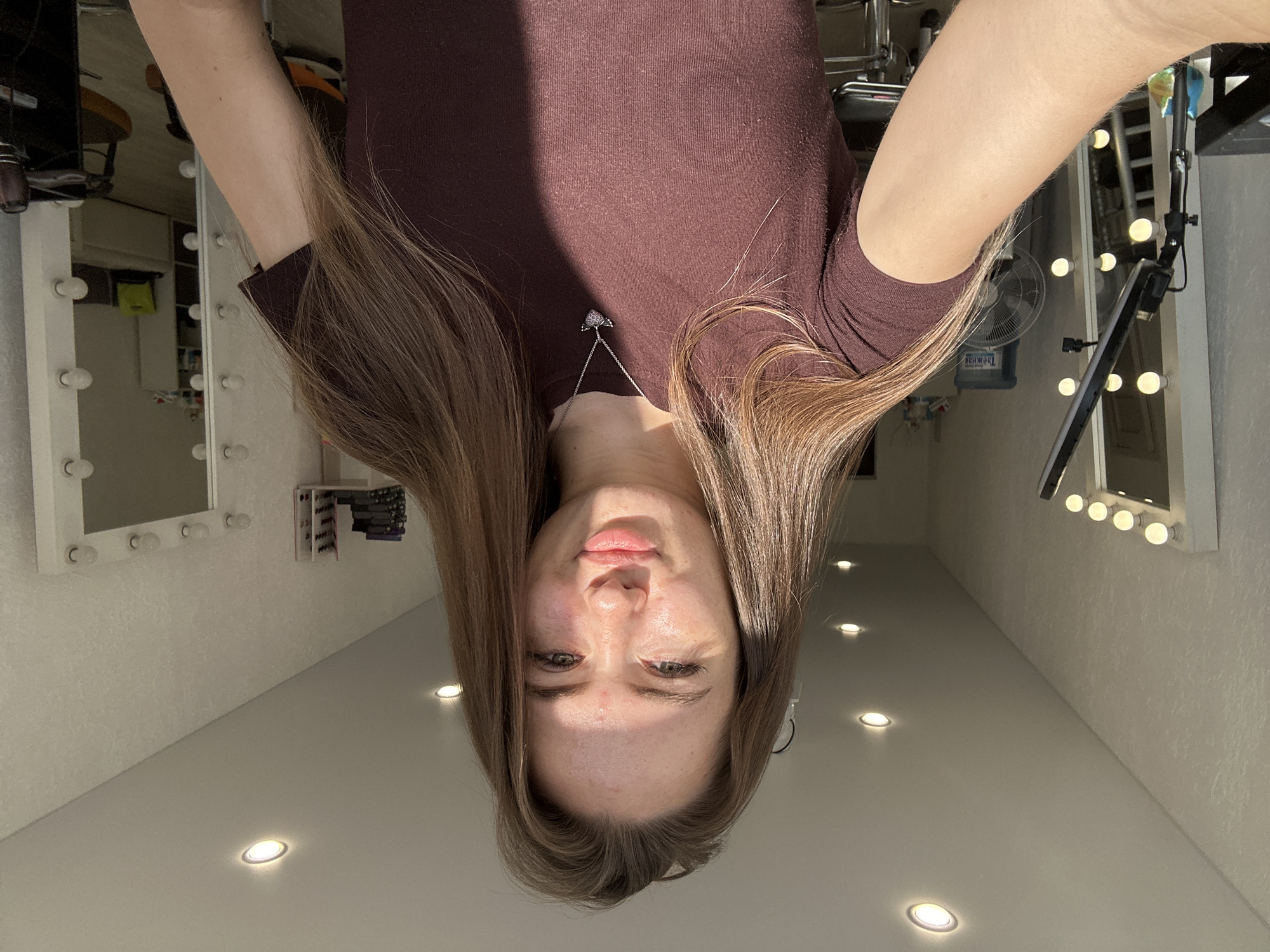 A young Caucasian woman with long, light brown hair is taking a selfie in a room that appears to be a makeup or dressing area. She is wearing a dark maroon t-shirt and a heart-shaped necklace with wings. The room has a white ceiling with recessed lighting and textured walls. To her left, a large mirror with multiple light bulbs is visible, along with a ring light stand. To her right, another large mirror with light bulbs reflects the room, showing shelves, a water cooler, a fan, and furniture. The woman is facing the camera, making a kissing gesture with her lips, and her right arm is extended, holding the camera. Sunlight is streaming in from the left, casting a shadow across her chest and the right side of the room.