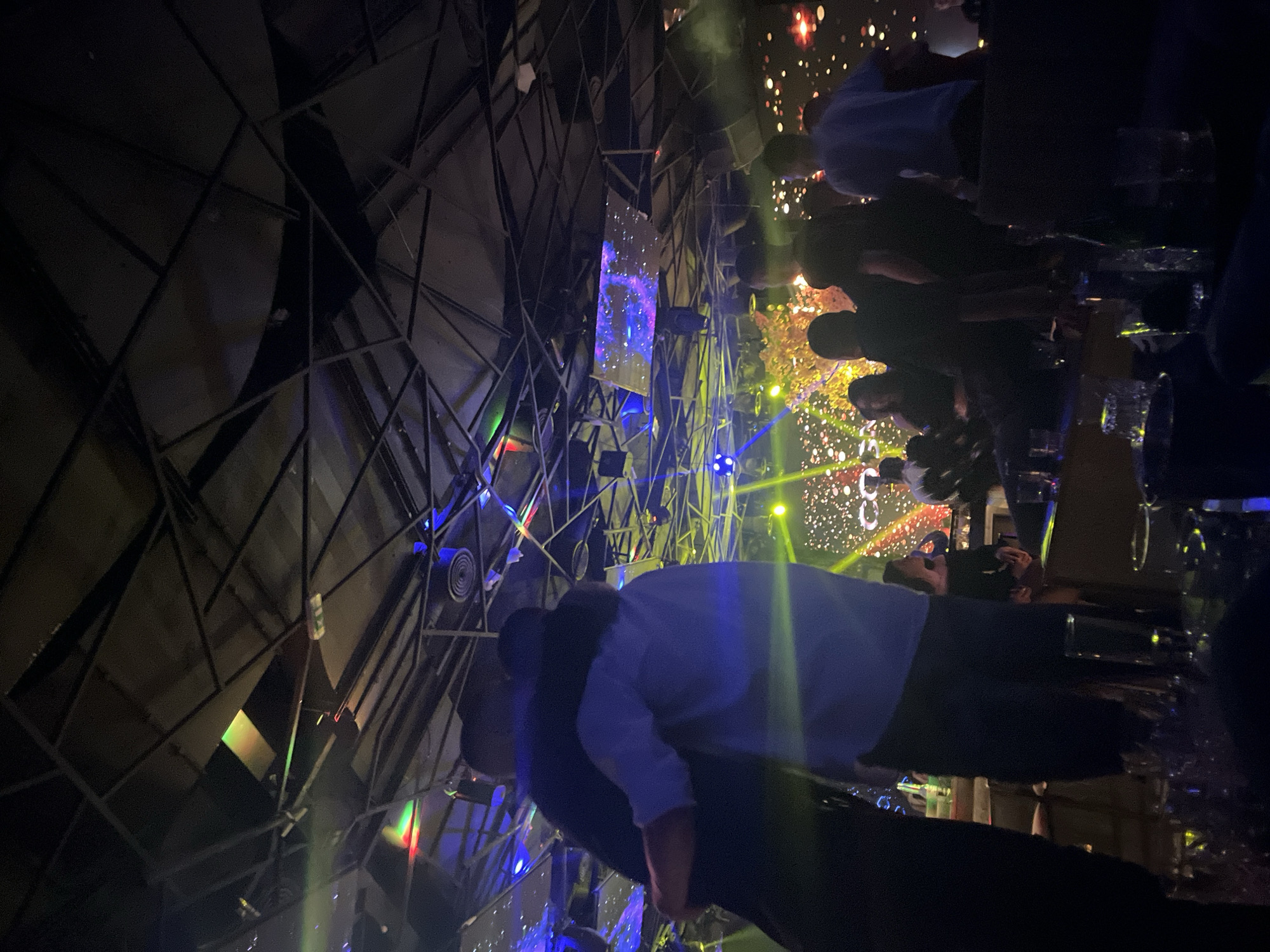 The image depicts the interior of a nightclub, possibly located in Plovdiv, Bulgaria, judging from the style of entertainment venues in the city. The lighting is dim, with laser beams and colored lights creating a dynamic atmosphere. A complex geometric structure of metal beams forms a cage-like element in the background. In the foreground and mid-ground, blurry figures of people suggest a crowded event. Some individuals are seen holding glasses and possibly taking photos with their phones. In the background, screens display images and possibly numbers, indicating a countdown or a live show. The scene shows a lively, urban nightlife environment.