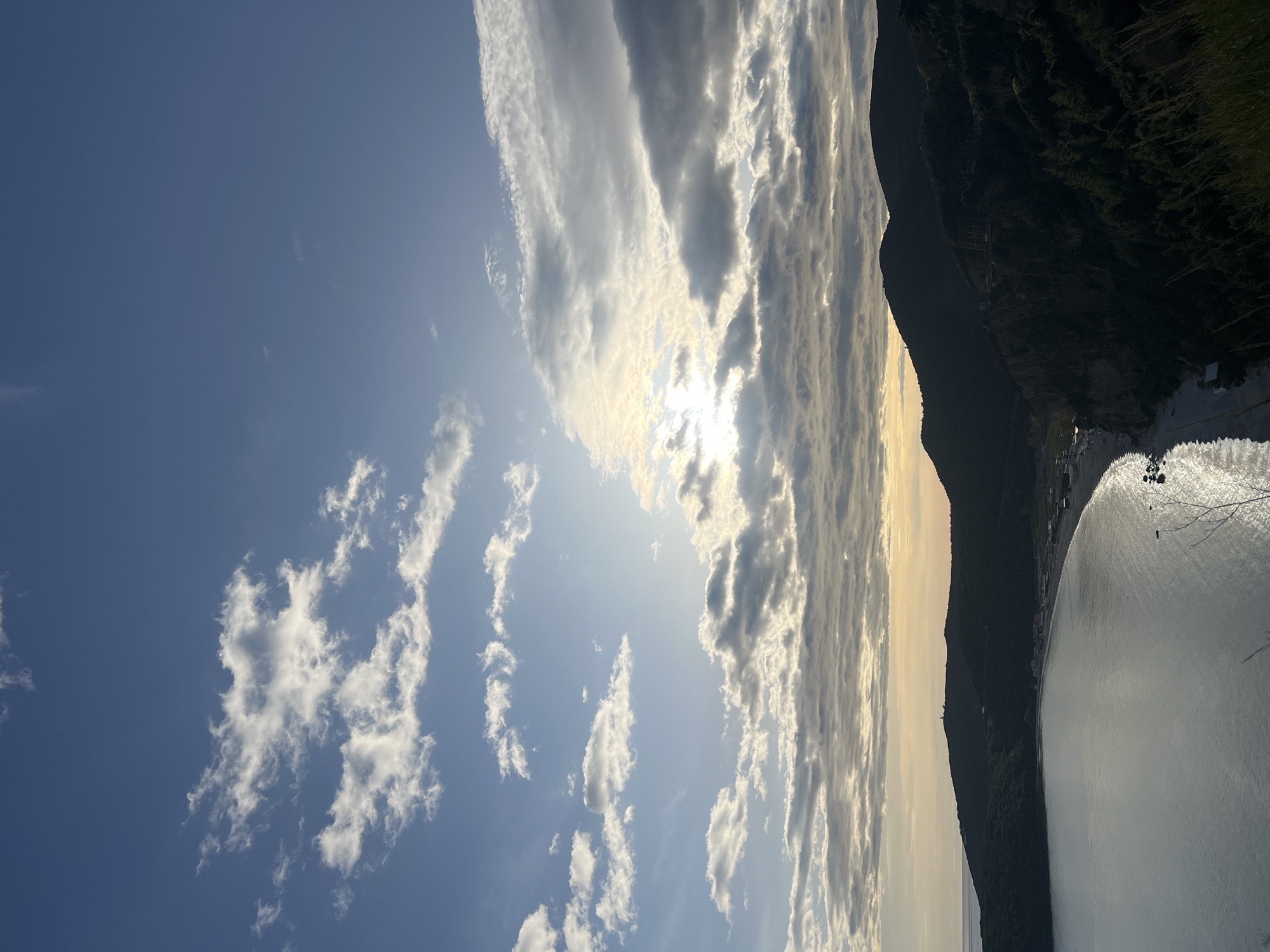 The image shows a coastline in Prijevor, Montenegro. A mountain ridge extends along the right side of the frame, bordering the edge of a body of water. A beach with a few buildings is nestled at the base of the mountain. The sky is filled with striated clouds with patches of clear blue. The sun is partially obscured by the clouds.