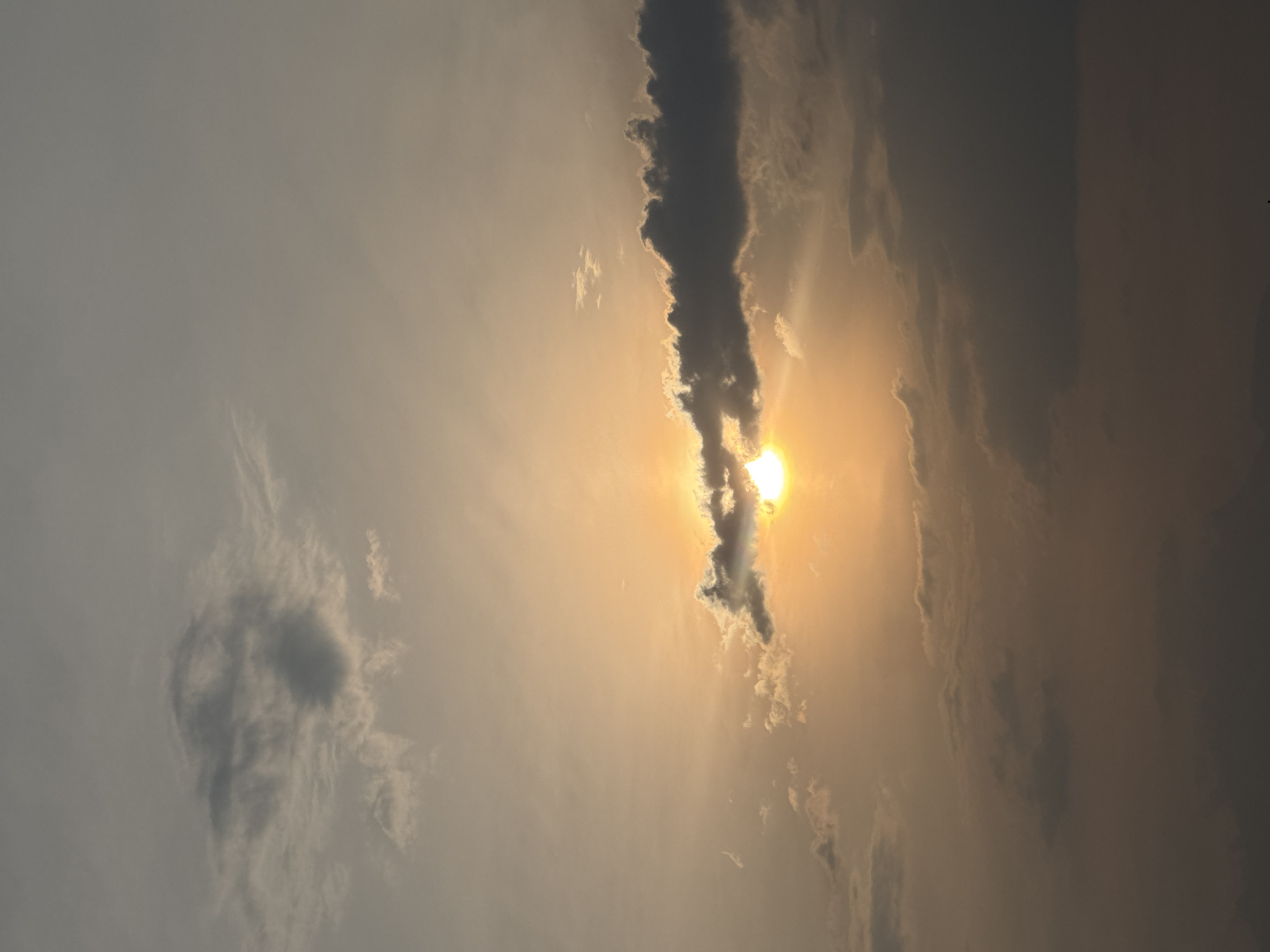 The image is a vertical shot of a sky scene, possibly taken in San Francisco Ocotlán (Ocotlán), Mexico. The sun is visible, partially obscured by a dark, vertical cloud formation that runs through the center of the frame. The sun's light is diffused and appears as a bright, golden orb, creating a warm glow in the surrounding sky. Other clouds are scattered throughout the sky, some with more defined shapes and some appearing as wispy streaks. The sky's color ranges from a light gray on the left to a darker, more muted tone on the right.