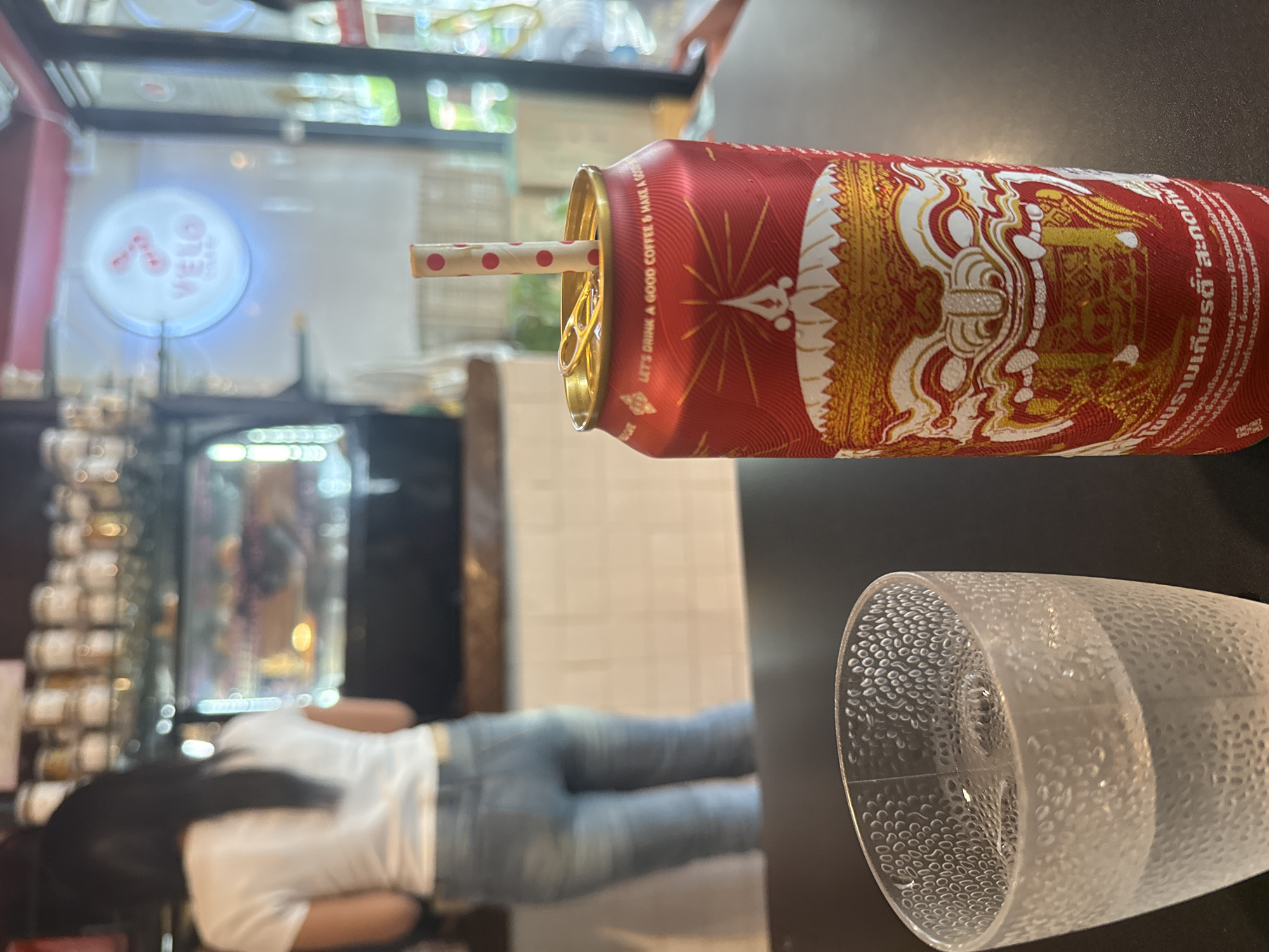 In Hua Hin City Municipality, Thailand, a red aluminum can with a distinctive mythical creature design and a pink-dotted straw is placed on a dark table. Next to it is a clear, textured glass filled with a clear liquid. In the background, a woman wearing a white t-shirt and light blue jeans is visible from the back, standing in front of a display case filled with food items. A circular neon sign with the word "VELO" is illuminated on the wall behind her. The overall setting appears to be a cafe or eatery.