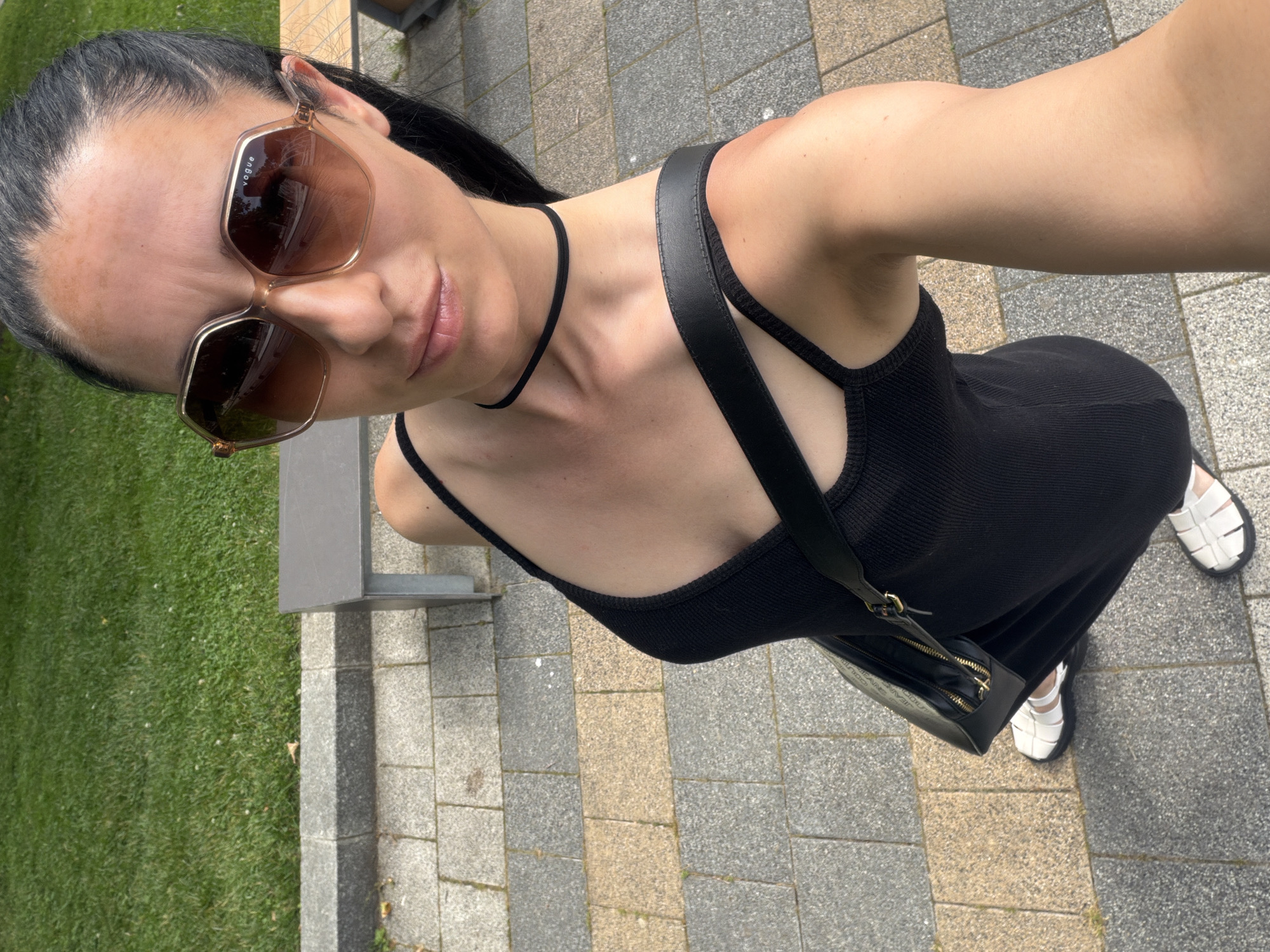 A woman stands on a paved area in Pleven, Bulgaria, with a grassy area and a bench visible in the background. She is wearing a black sleeveless dress, white sandals, sunglasses, and a black choker necklace. A black purse with a gold zipper is slung over her shoulder. The woman is looking at the camera and holding it out to take a selfie. The lighting suggests it is daytime.