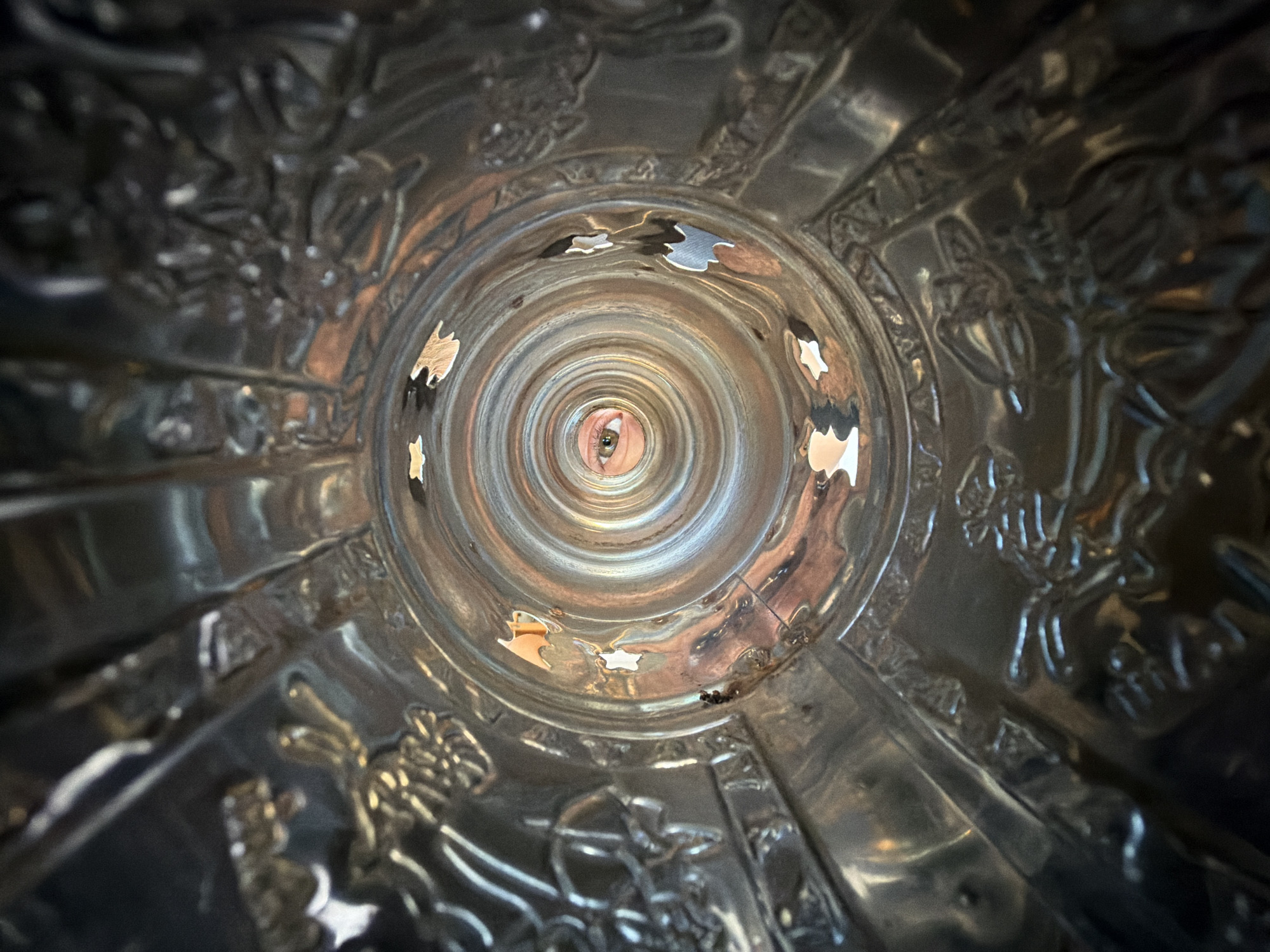 The image is a close-up, upward-looking shot from inside a decorative metallic object, possibly a vase or a bowl, with intricate relief carvings on its inner surface. The surface is highly reflective, showing concentric rings and distorted reflections of the surroundings. A single human eye, with a green iris, is visible through a circular opening at the deepest point of the object. The eye appears to be looking directly at the camera. Scattered around the opening are small, irregularly shaped pieces of what appear to be puzzle pieces, some reflecting light and others appearing darker. The lighting suggests an indoor environment. No specific location in Chaikovsky, Russia, can be determined from the visual information in the image.