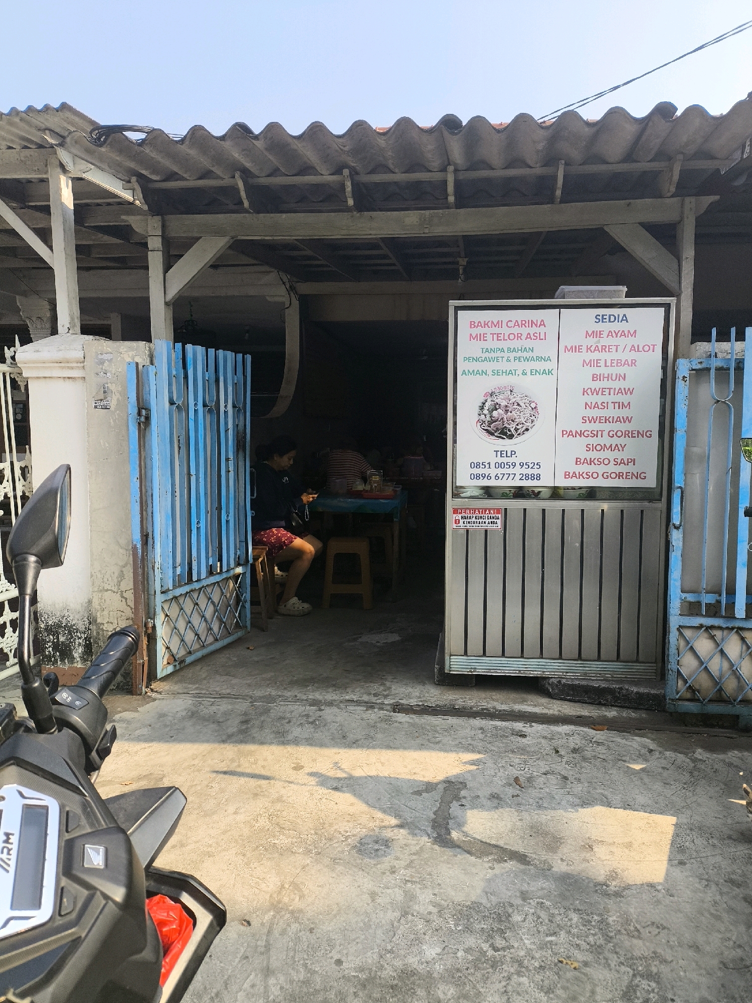 A front exterior view shows a small food stall located in West Jakarta, Indonesia, specializing in noodles and other dishes. The stall has a corrugated roof supported by weathered wooden beams. A bright blue metal gate, partially open, provides access to the interior, where a person is seated at a table, engrossed in their phone. Another person is visible in the background. The stall's menu is displayed on a sign, advertising "Bakmi Carina Mie Telor Asli" and various noodle dishes, as well as "Pangsit Goreng," "Siomay," "Bakso Sapi," and "Bakso Goreng." The ground is paved with concrete and shows signs of wear and tear, with shadows cast by the sunlight. A motorcycle is parked in the foreground, its handlebars and part of the front panel visible.