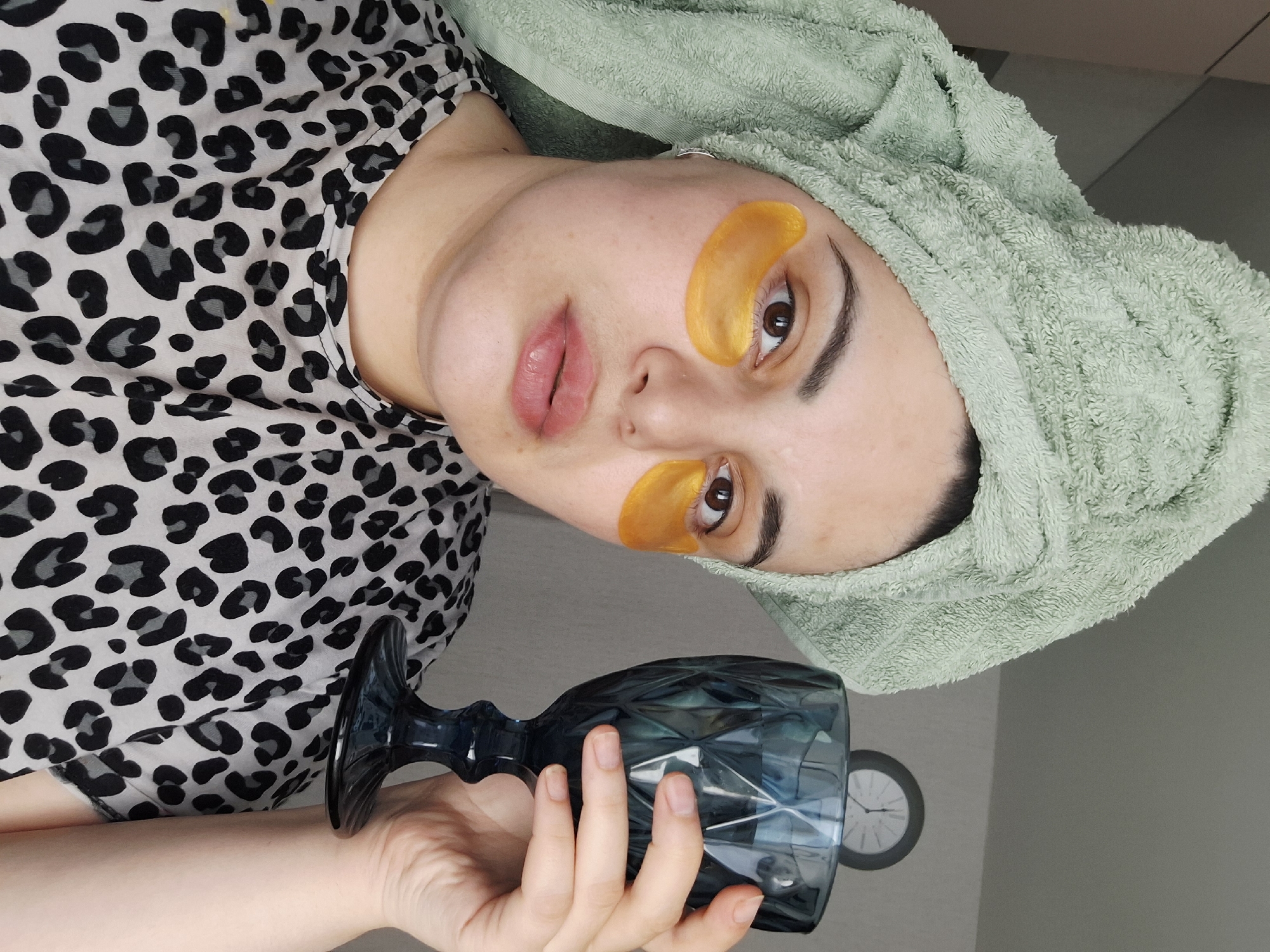 A young woman is posing indoors. She has a green towel wrapped around her head and gold eye masks under her eyes. She is wearing a black and white animal print top. She holds a textured blue glass in her right hand. Behind her is a grey wall, with a white clock visible in the background. The location is specified as the city of Амзя, Russia.