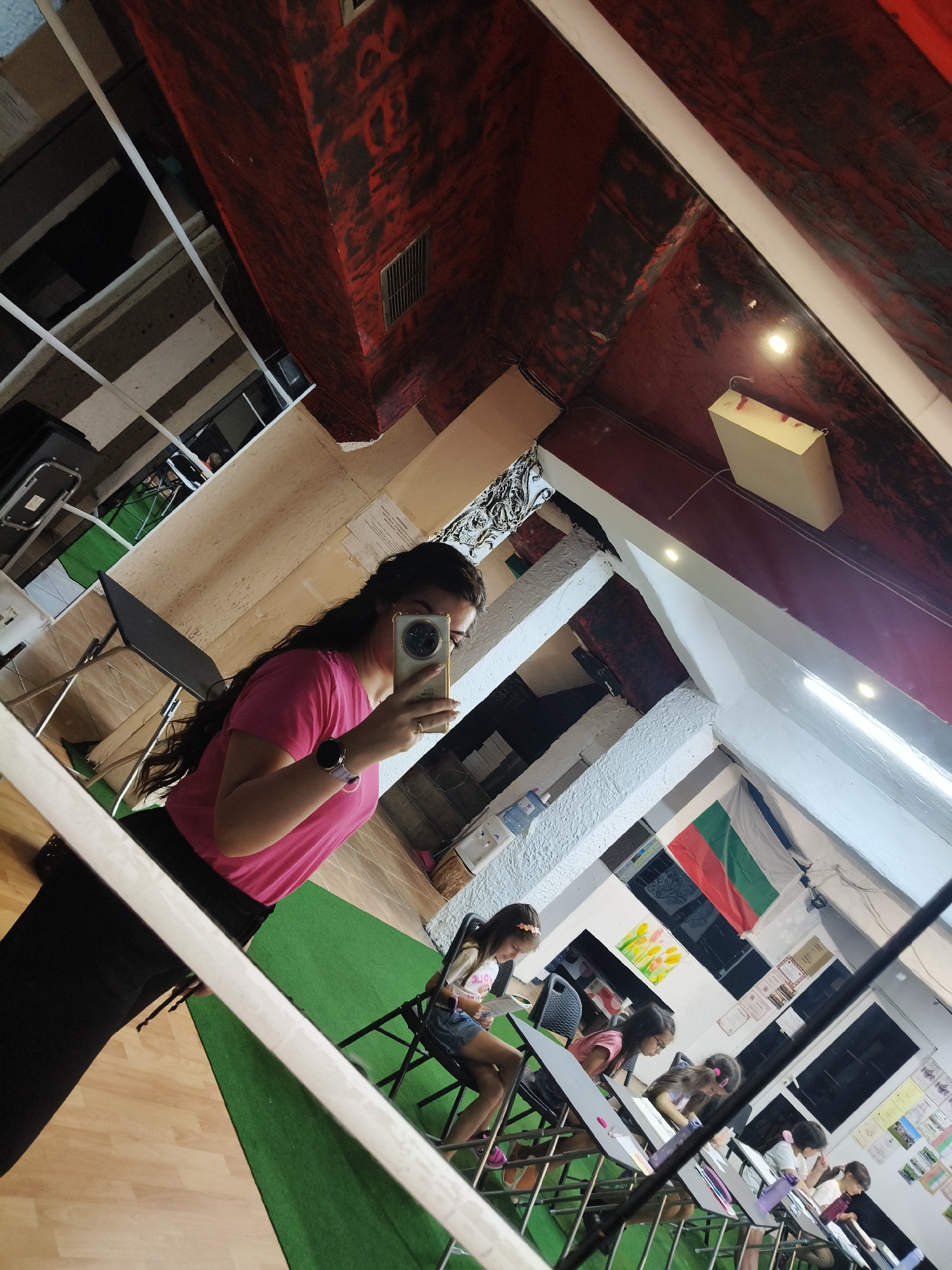 A woman is taking a selfie in a room with a red textured ceiling and white beams. In the background, several young girls are sitting at desks, appearing to be engaged in an art class. The room has a green carpeted area and wooden flooring. A Bulgarian flag hangs on the wall. The overall impression is that of an art studio or classroom in Plovdiv, Bulgaria.