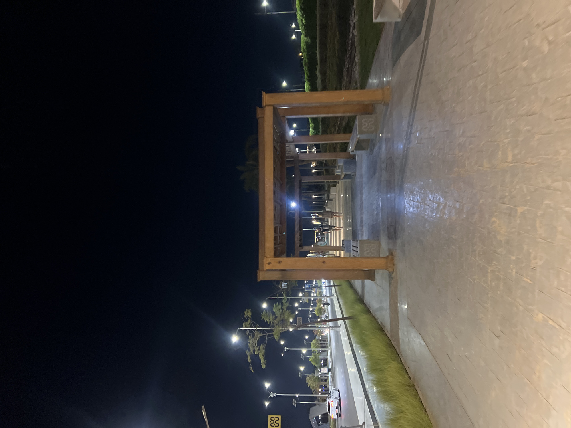 This is a nighttime exterior, eye-level, medium shot of a promenade in Sharm El Sheikh, Egypt. A wooden pergola structure with benches is centrally located, with a paved walkway leading through it. Two individuals, a man and a woman, are walking away from the camera within the pergola. To the left of the walkway is a strip of green grass and then a street with a white car driving away. Multiple streetlights illuminate the scene, creating a dark blue sky with a few bright lights. The ground appears wet and reflective, possibly from recent rain. The overall atmosphere is calm and serene.