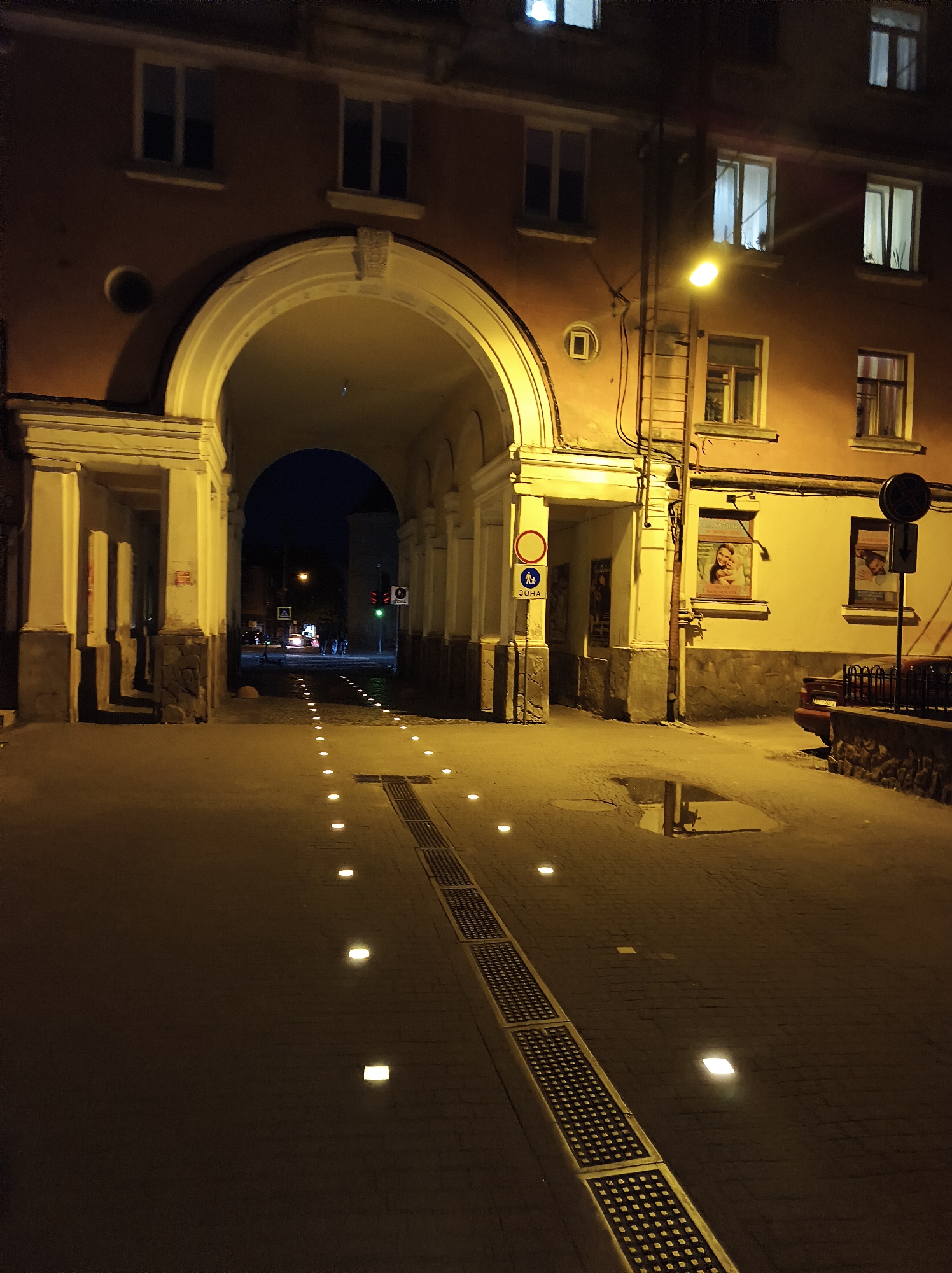 The image depicts an archway leading into a courtyard or street at night in Ternopil, Ukraine. The building facade is a warm orange hue. Several windows are visible, some illuminated from within. A streetlamp casts a yellow glow on the scene.
The ground is paved with cobblestones, and a series of small, square lights are embedded along a path leading through the archway. A drainage grate runs alongside these lights. In the distance, beyond the arch, car headlights are visible, along with the silhouettes of people and a traffic light showing green. A sign with a pedestrian symbol and the word "ЗОНА" (Zone) is posted on the left. A puddle reflects the surrounding lights on the right side of the pathway. A dark-colored car is parked on the right. Posters are displayed on the building's wall within the archway. The overall atmosphere is dimly lit and quiet, suggesting a late hour.