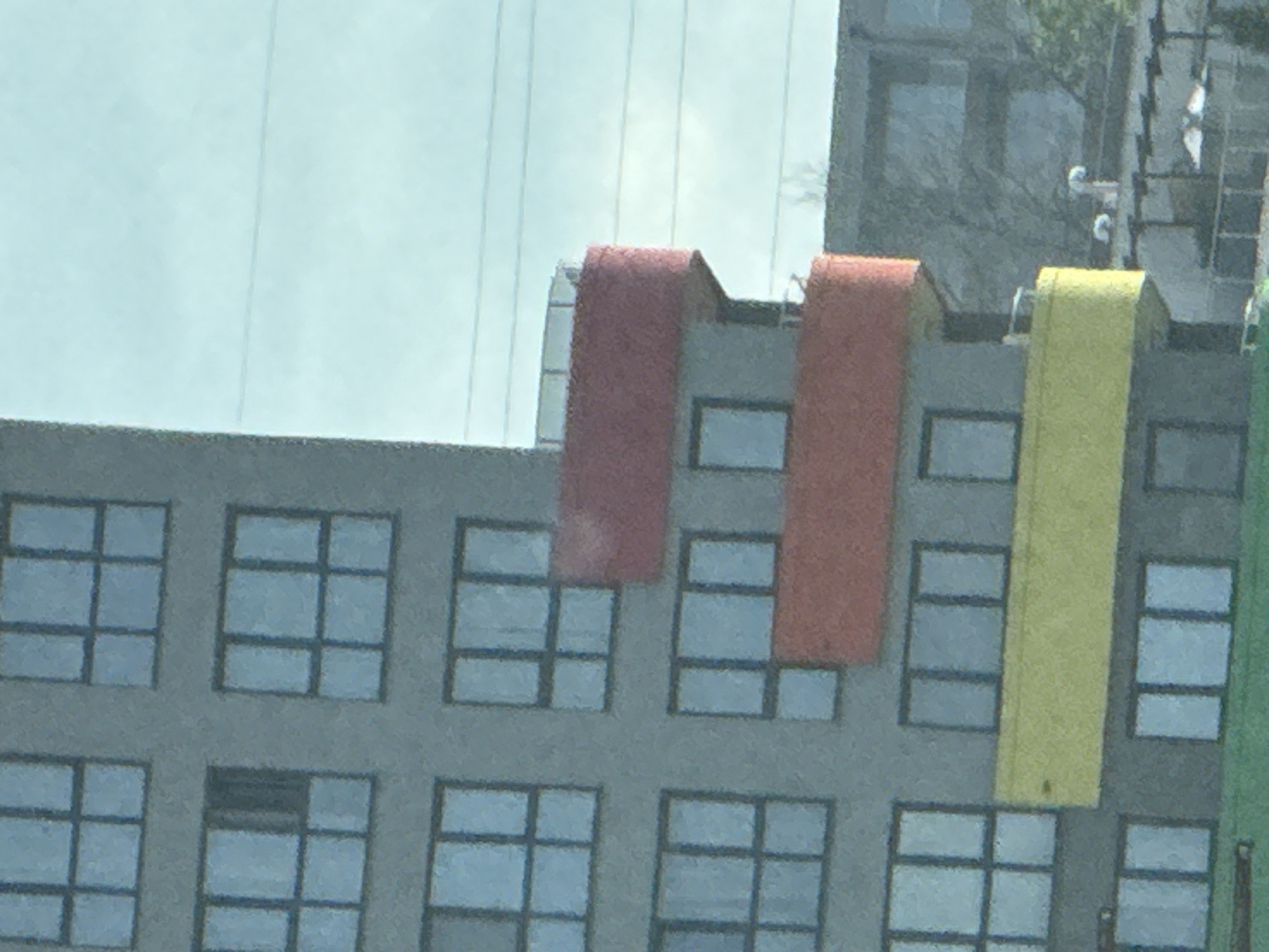 The image shows a multi-story building with a grey facade and numerous windows. The building has balconies with colored panels in red and yellow. The red panels are on the upper floors, and the yellow panel is on a lower floor. The windows appear to be modern, with dark frames and multiple panes. In the background, another building with a darker facade is visible, also with windows. Electrical wires are stretched across the sky. The scene is likely urban, given the architecture and the presence of power lines.