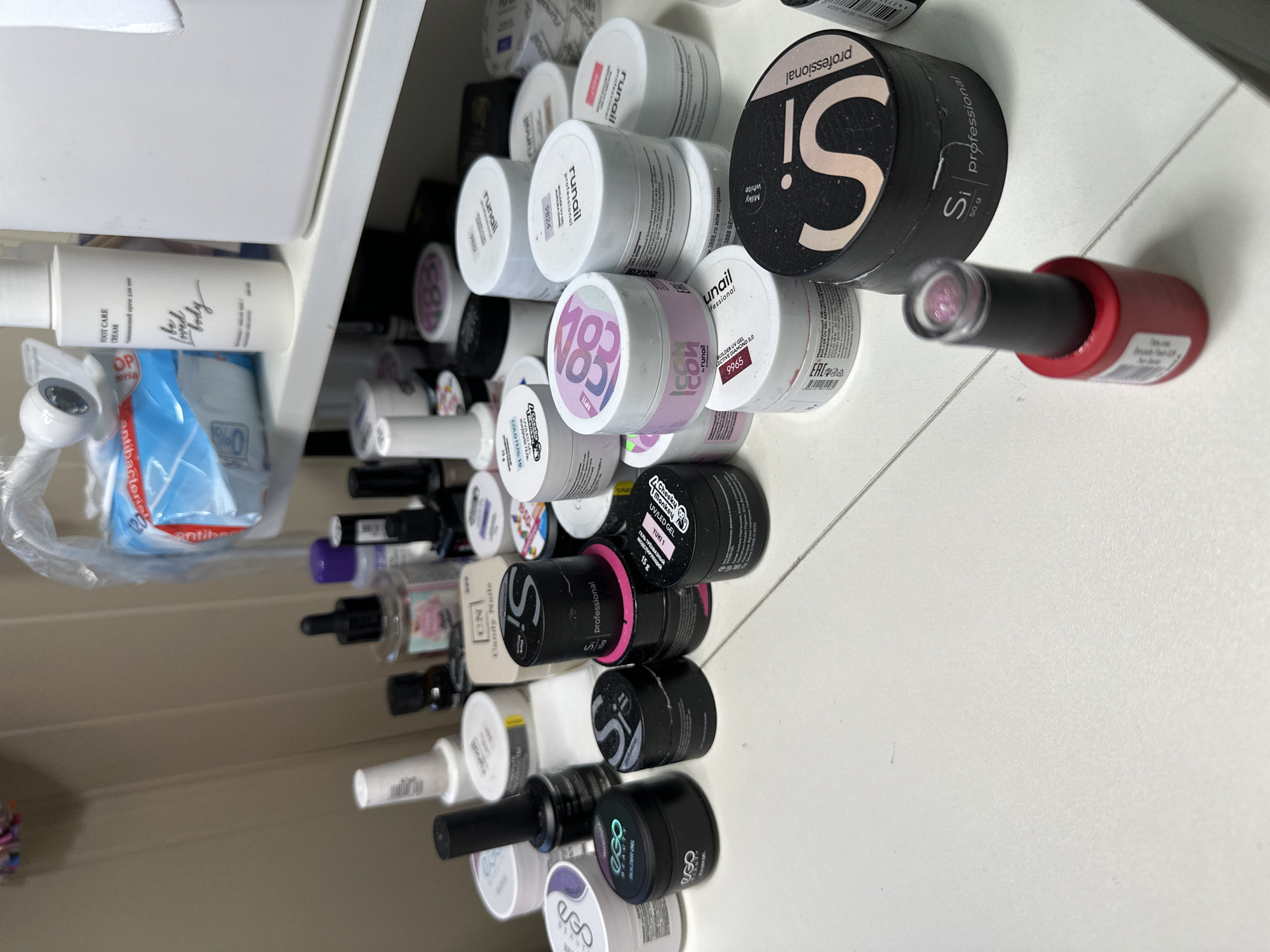 The image depicts a collection of nail care products arranged on a white surface, likely a table or shelf. The products include various containers of gels, polishes, and creams, many of which are labeled with brand names such as "Si Professional," "Runail," and "Eco Beauty." There are also some bottles of liquid products and a packet of wipes. The overall impression is one of a personal collection or a small business inventory. The scene appears to be indoors, with neutral lighting and a plain background. The exact location within Novoaltaysk, Russia, cannot be determined from the visual information provided.