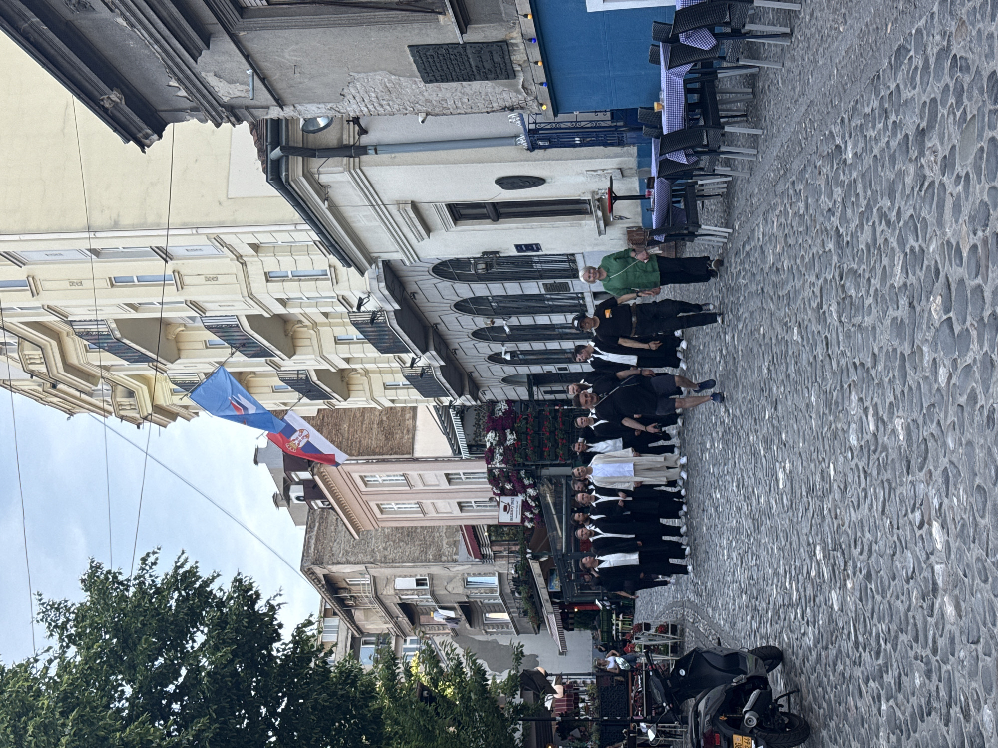 The image captures a scene on a cobbled street in Belgrade, Serbia. A group of people, mostly dressed in black suits with some in white shirts, are lined up along the street. A Serbian flag hangs between buildings on the left side of the frame. In the background, a restaurant with blue accents is visible. Several parked motorcycles are at the bottom of the frame. The environment includes buildings of various heights and architectural styles lining both sides of the narrow street, with trees visible in the lower-left corner.