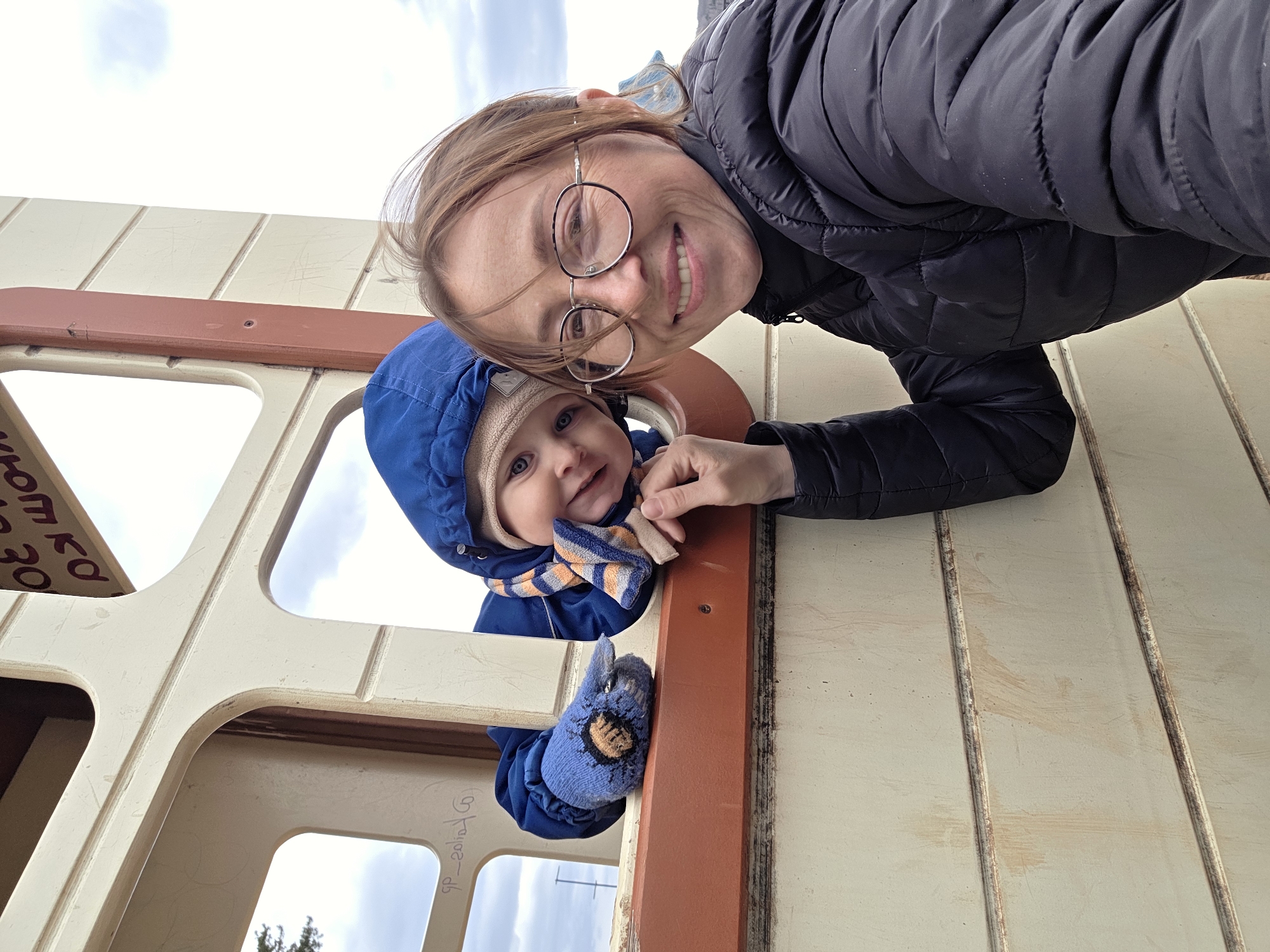 The vertical portrait captures a cheerful moment between an adult woman and a baby or toddler within a playground structure in Chaikovsky, Russia. The woman, positioned at the upper right, has light brown hair, wears glasses with dark frames, and a dark, possibly black, puffer jacket. She is smiling broadly, looking directly at the camera.
The baby, located in the lower-middle left, is inside the playground structure, peeking out from an opening. The baby has bright blue eyes and is dressed warmly in a blue hooded jacket, a light brown knitted hat, and a striped scarf featuring orange, blue, and beige colors. A blue mitten with an orange and brown design is visible on one of the baby's hands, resting on the wooden frame. The baby is also smiling, engaging with the camera.
The setting appears to be an outdoor children's playground, characterized by a sturdy, light-colored wooden or plastic play structure with darker brown trim. The sky, visible through the various openings and reflections, is bright but appears overcast, suggesting cool or cold weather, consistent with the heavy clothing worn by both individuals. The overall mood is joyful and affectionate.
Visible text on the left side, reflected upside down in one of the window-like openings, reads "от 3 до 30", which translates to "from 3 to 30", possibly indicating an age range for the playground equipment. Another partially visible, upside-down reflection on the right appears to be a social media handle, "@_fairos_db".
