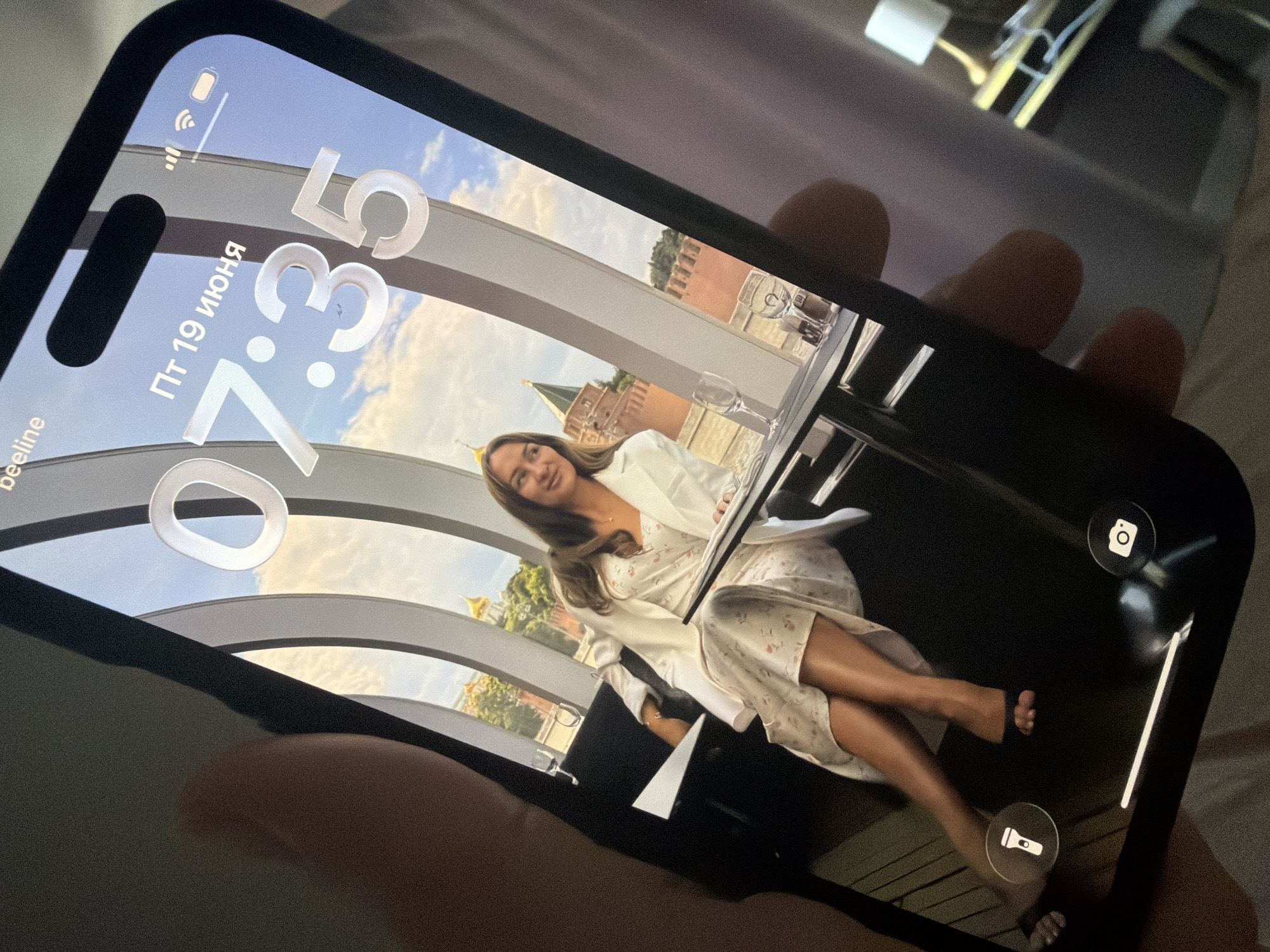 A woman is seated at a table in front of a large window with a city view. She is wearing a white blazer over a floral dress. The window offers a view of a red brick building with green domes, possibly a historical landmark in Malinovka, Russia. The time displayed on the phone screen is 7:35 and the date is June 19th. The phone is being held by a hand with visible fingers and thumb, partially obscuring the left side of the image. The environment appears to be an indoor seating area, possibly a restaurant or cafe, with additional tables and chairs visible. The lighting is dim, with a lamp visible in the background.