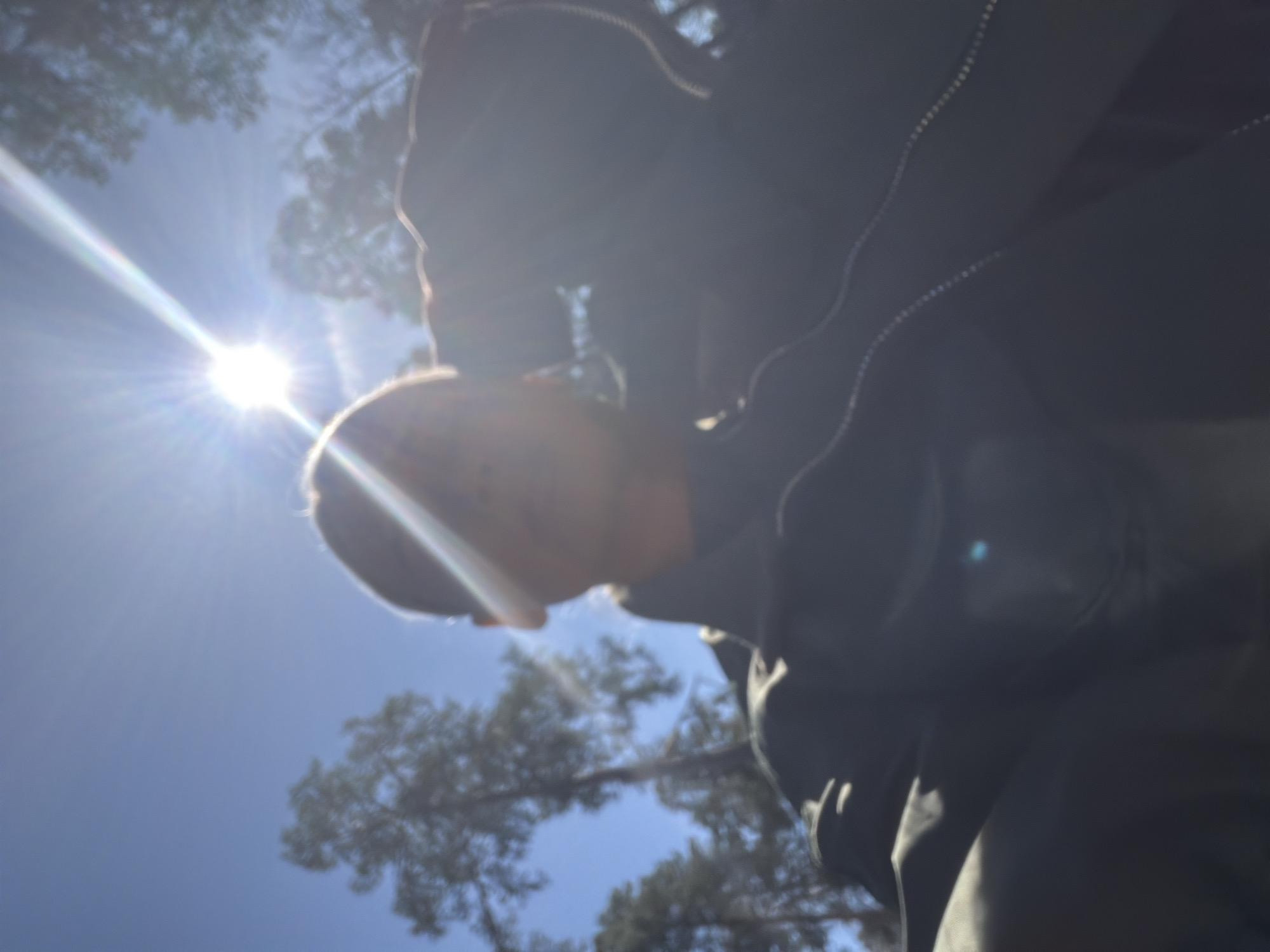 The image shows a low-angle view of a person looking downward towards the ground in an outdoor setting. The bright sun is visible in the upper left of the frame, causing significant lens flare. The person is wearing a dark-colored jacket with visible stitching details. Trees are visible in the background, silhouetted against the blue sky. It is not possible to determine the location within Sarapul, Russia from this image alone.