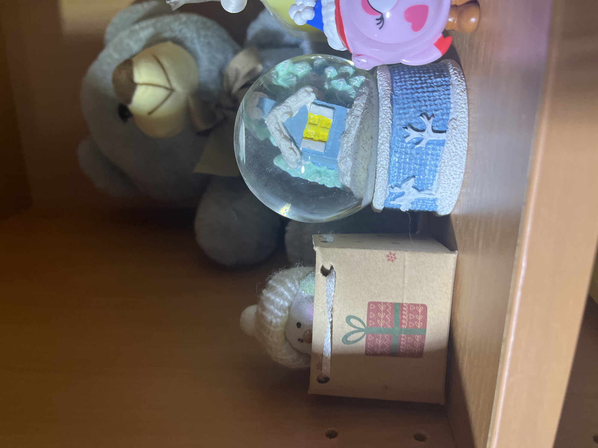 The image shows a shelf with several decorative items. On the left is a small brown paper gift bag with a red and green present design, and a white fuzzy object, possibly a toy or figurine, peeking out of the top wearing a white pom-pom hat. To the right of the gift bag is a snow globe with a blue house and snow-covered trees inside, set on a blue and white textured base. Further to the right, a portion of a pink owl-shaped figurine with a blue and red hat is visible. In the background, a large, light blue teddy bear is partially visible. The shelf appears to be made of wood, and the background is also wooden. There is no indication of the city of Ashkadar, Russia, or any specific location within it. The image is a still shot and depicts inanimate objects arranged on a shelf.