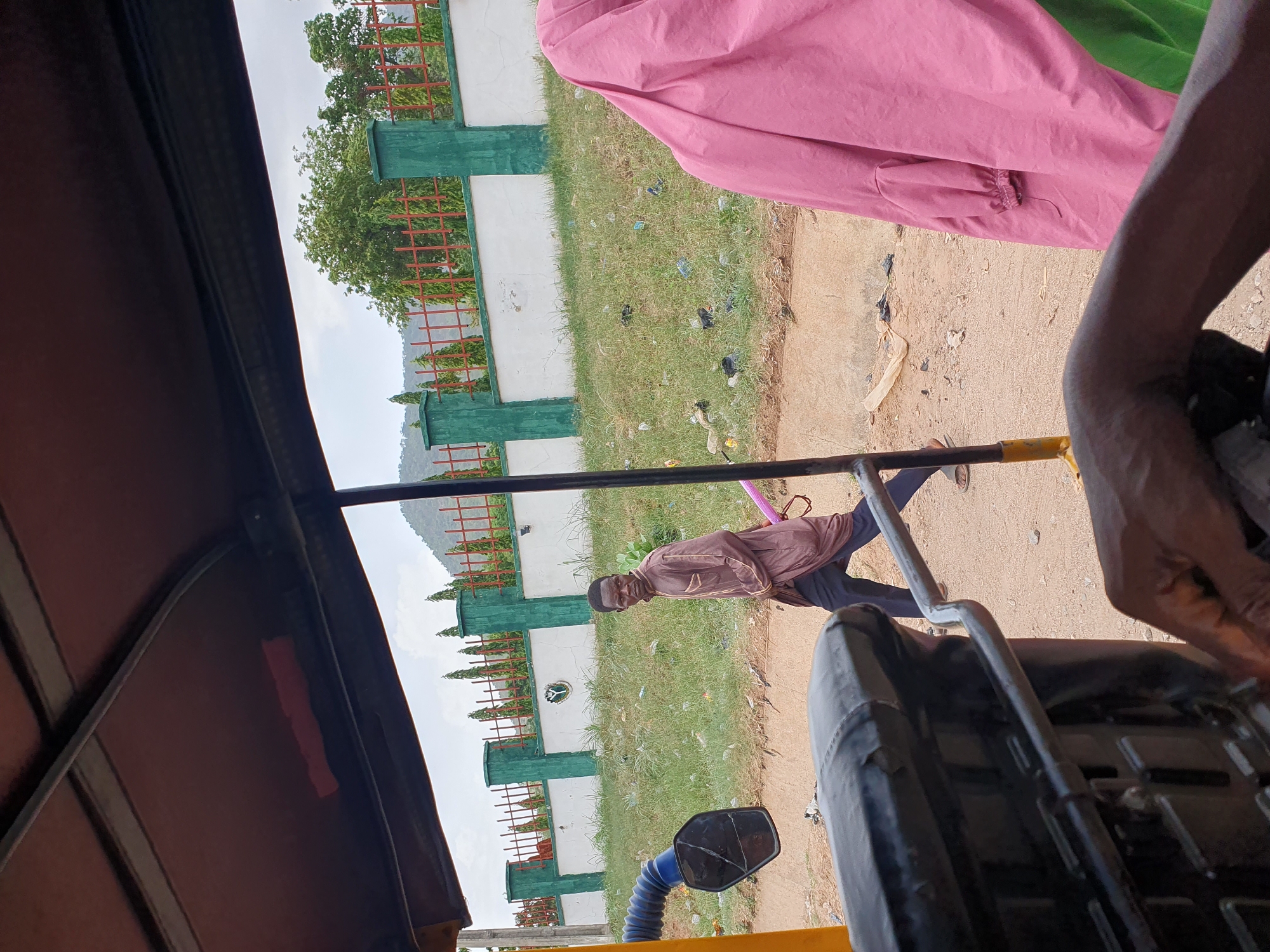 The image shows a street scene in Lokoja, Nigeria, viewed from inside a vehicle, likely a yellow commercial tricycle (keke). An adult male wearing a brown shirt and dark trousers stands on the roadside, facing towards the vehicle. Behind him is a white concrete wall with green painted pillars and red metal fencing on top. Grass grows between the wall and the road. Scattered trash is visible on the ground. Parts of the vehicle, including the seat and handlebars, are visible in the foreground. Other passengers, partially obscured, are wearing pink and green clothing. The sky is overcast.