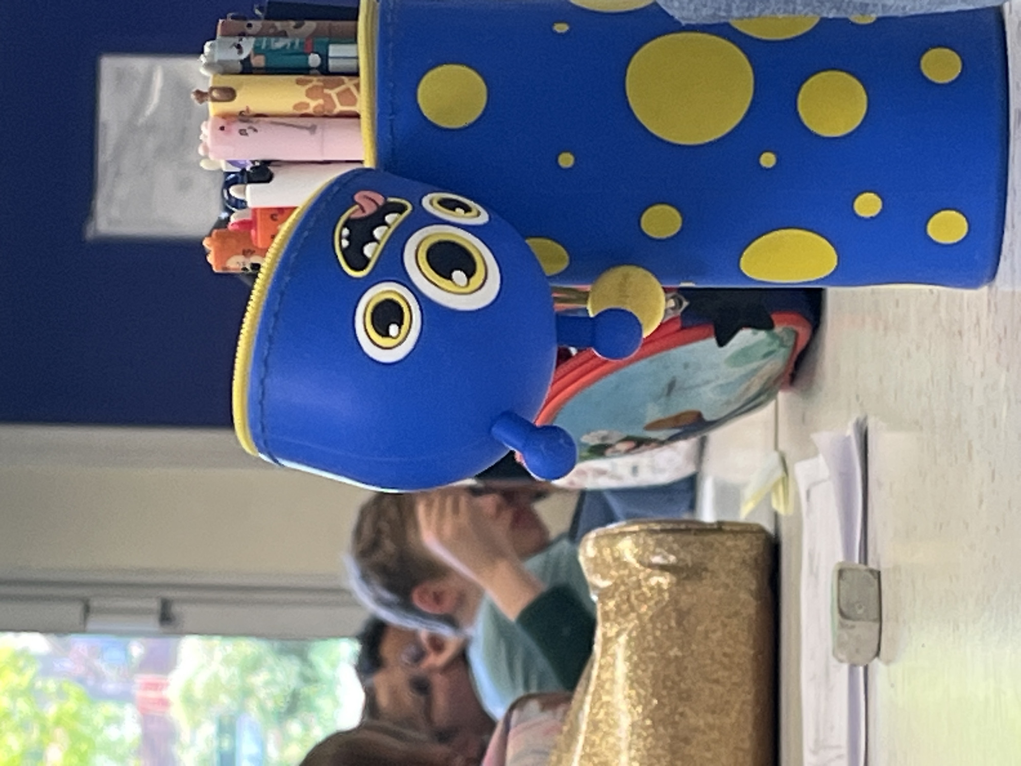 The image, taken in what appears to be an indoor setting like a classroom or study area, captures a close-up of stationery items on a table or desk, with blurred figures in the background.
The main subjects in the foreground are two blue pencil cases. The larger one, positioned upright, is cylindrical with a zipper and features a pattern of scattered yellow polka dots. Partially visible inside it are several pens or pencils topped with small, animal-shaped figures, including a giraffe, a pink pig, and a fox. Next to it is a smaller, character-shaped blue pencil case with exaggerated features, including large yellow-and-white eyes, a black and white open mouth with a pink tongue, and small blue, bulbous appendages. It also has a zipper, suggesting it can open.
In the mid-ground, a golden, textured bottle or container stands on the table to the right. Behind the stationery, two individuals are visible but heavily blurred. One person, with light brown hair, appears to be looking down or resting their hand near their face, wearing a green top. Another person is dimly visible behind them.
The background includes a window or light source that reveals blurred greenery and possibly structures outside, indicating it is daytime. A light-colored wall or surface with a paper clipped to it is visible on the far right. No legible text is visible anywhere in the image. The scene suggests a relaxed, everyday environment, possibly during a school day or a study session in La Fouillouse, France.