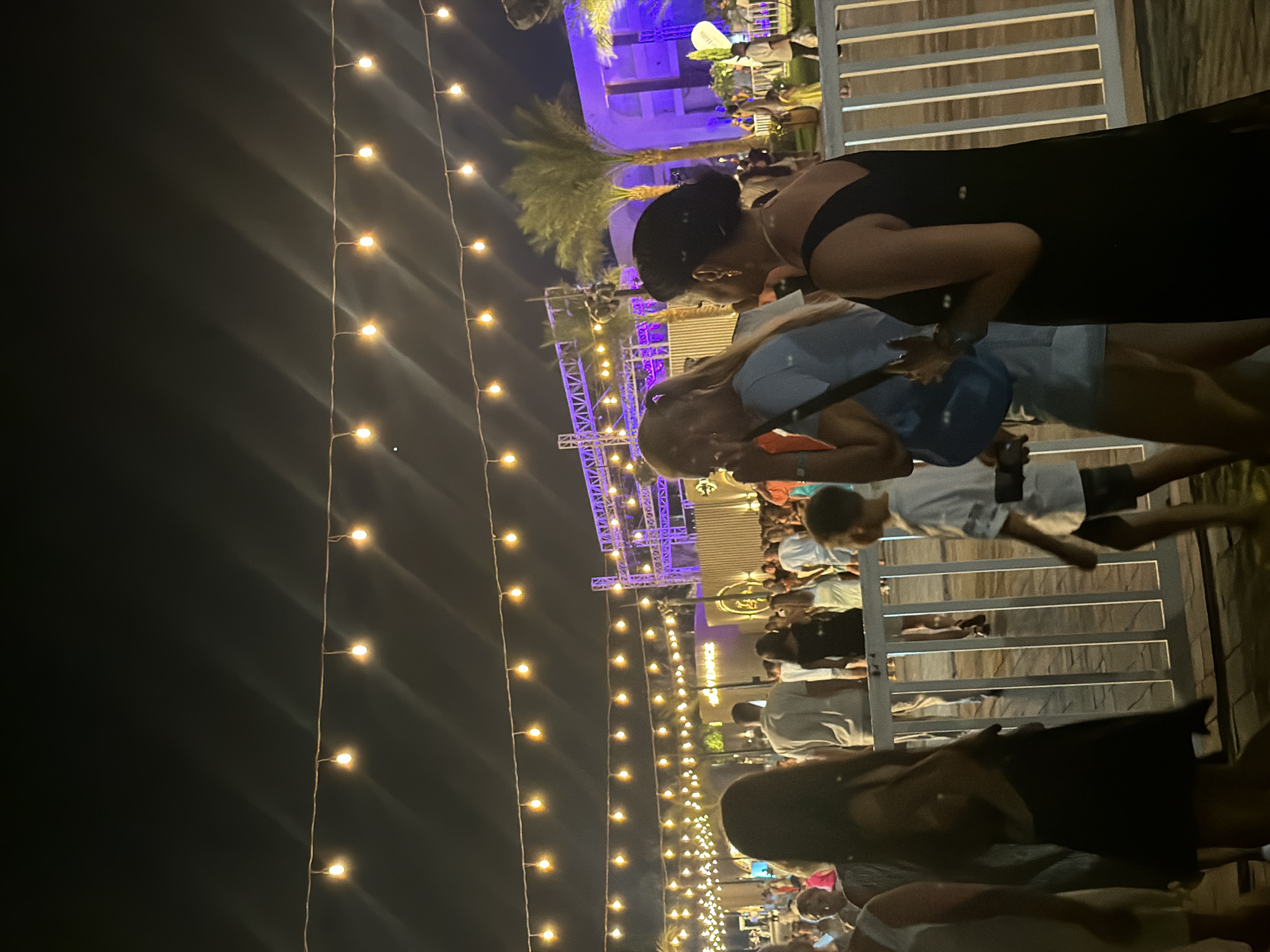 The image depicts an outdoor evening scene in Sharm El Sheikh, Egypt, likely at a beachfront venue or entertainment area, possibly at an event or festival. Several strings of warm-toned festoon lights are strung across the upper portion of the frame, casting a glow against the dark night sky. In the mid-ground, a crowd of people are visible, some standing and conversing, others walking. A stage structure with purple lighting is visible in the background, suggesting a performance or event is taking place. Palm trees are interspersed throughout the scene. A white metal fence runs along the lower part of the image, separating the viewer from the area where the people are gathered. The overall atmosphere appears festive and social.