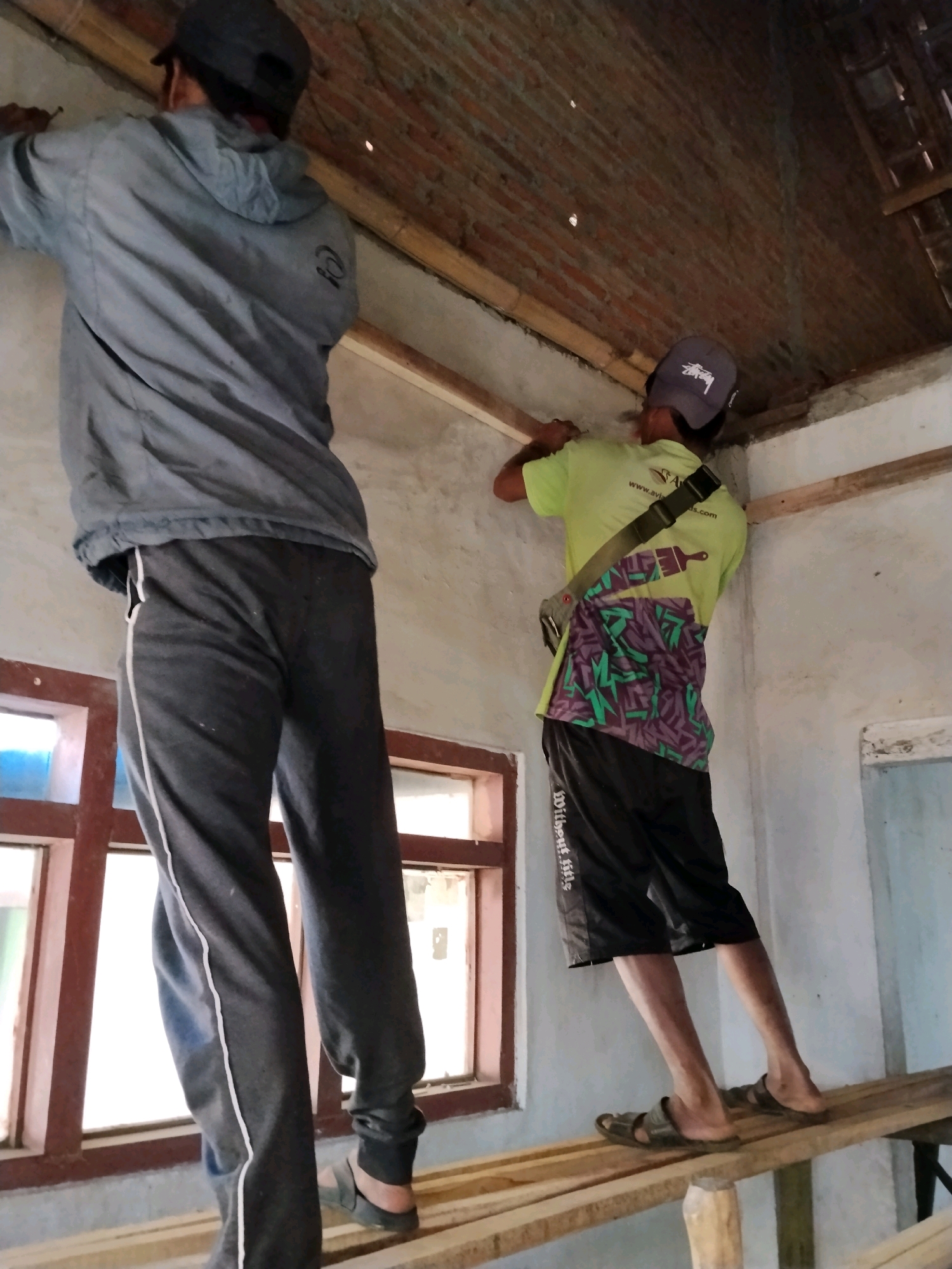 Two men are performing construction work on the interior of a building. The man on the left is wearing a dark cap, a light grey hooded jacket, and dark grey sweatpants with a white stripe down the side. He is standing on a wooden plank and appears to be working on the wall. The man on the right is wearing a dark cap with a white logo, a bright yellow t-shirt with a colorful design on the back, and black shorts. He is also standing on a wooden plank, higher up than the other man, and is working on the ceiling. Both men are using tools. The environment appears to be a partially constructed room with a brick ceiling and a window with reddish-brown wooden frames. Wooden beams are visible along the ceiling and forming part of the structure. The scene suggests ongoing renovation or construction activity. This image may have been taken in Panggungan, Indonesia, given the context provided.