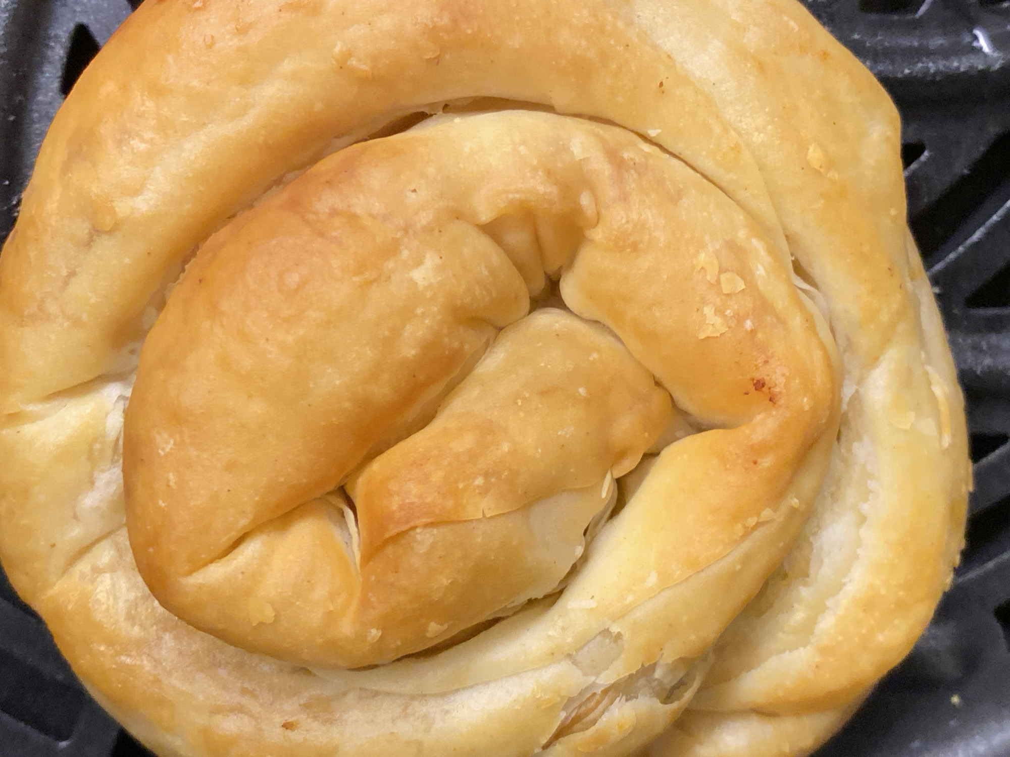 A close-up photograph captures a golden-brown, swirled pastry. It is likely a burek, a type of baked filled pastry made of thin flaky dough, common in the Balkans. The pastry exhibits a layered texture, with the dough tightly coiled into a circular shape. No specific location in Budva, Montenegro, is discernable from the image.