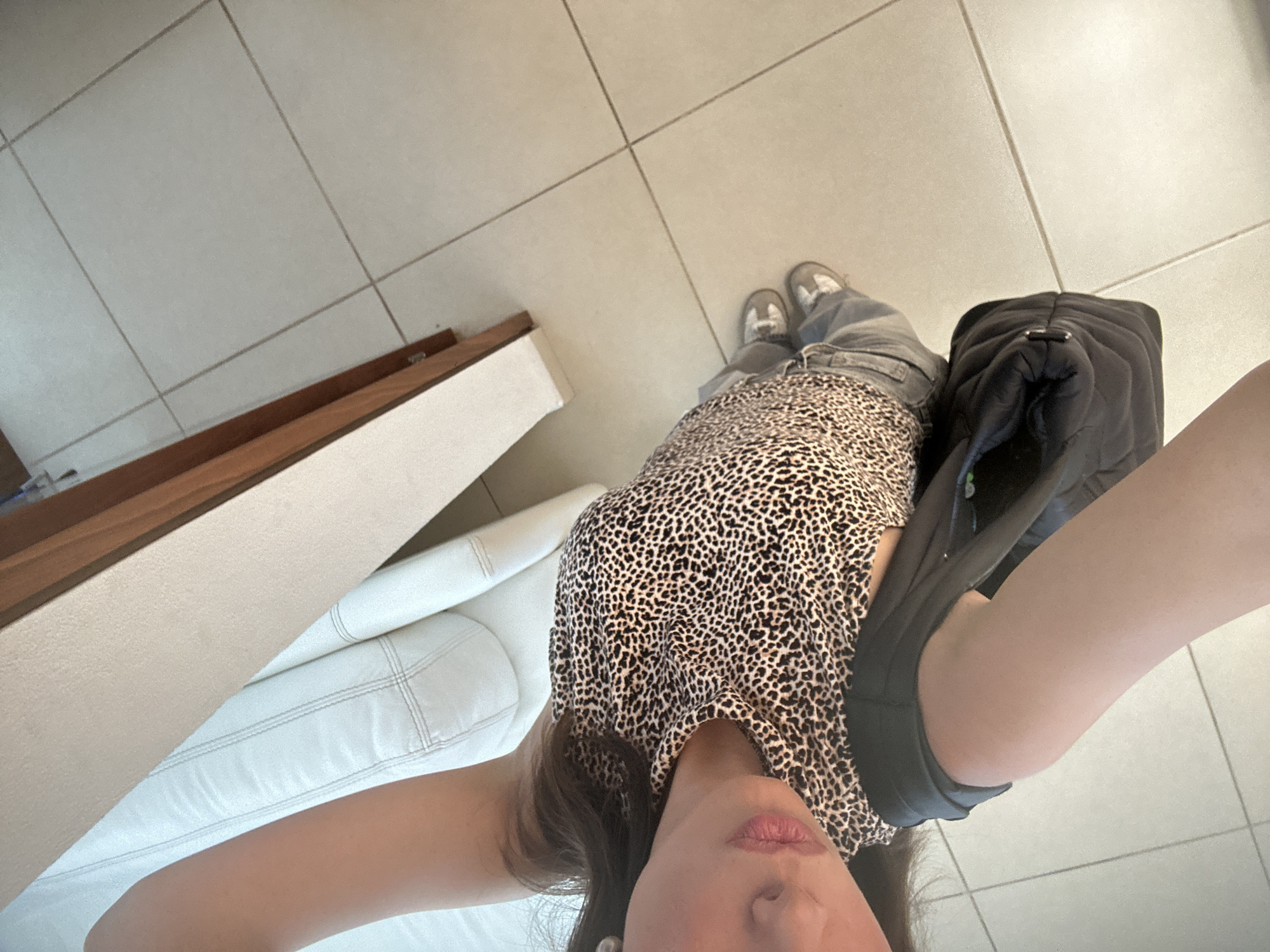 The image shows a person wearing a cheetah-print shirt, grey jeans, and grey shoes. They are standing on a tiled floor with a white leather sofa visible to the side. A black bag is slung over their shoulder. The location is Unknown, Unknown.
