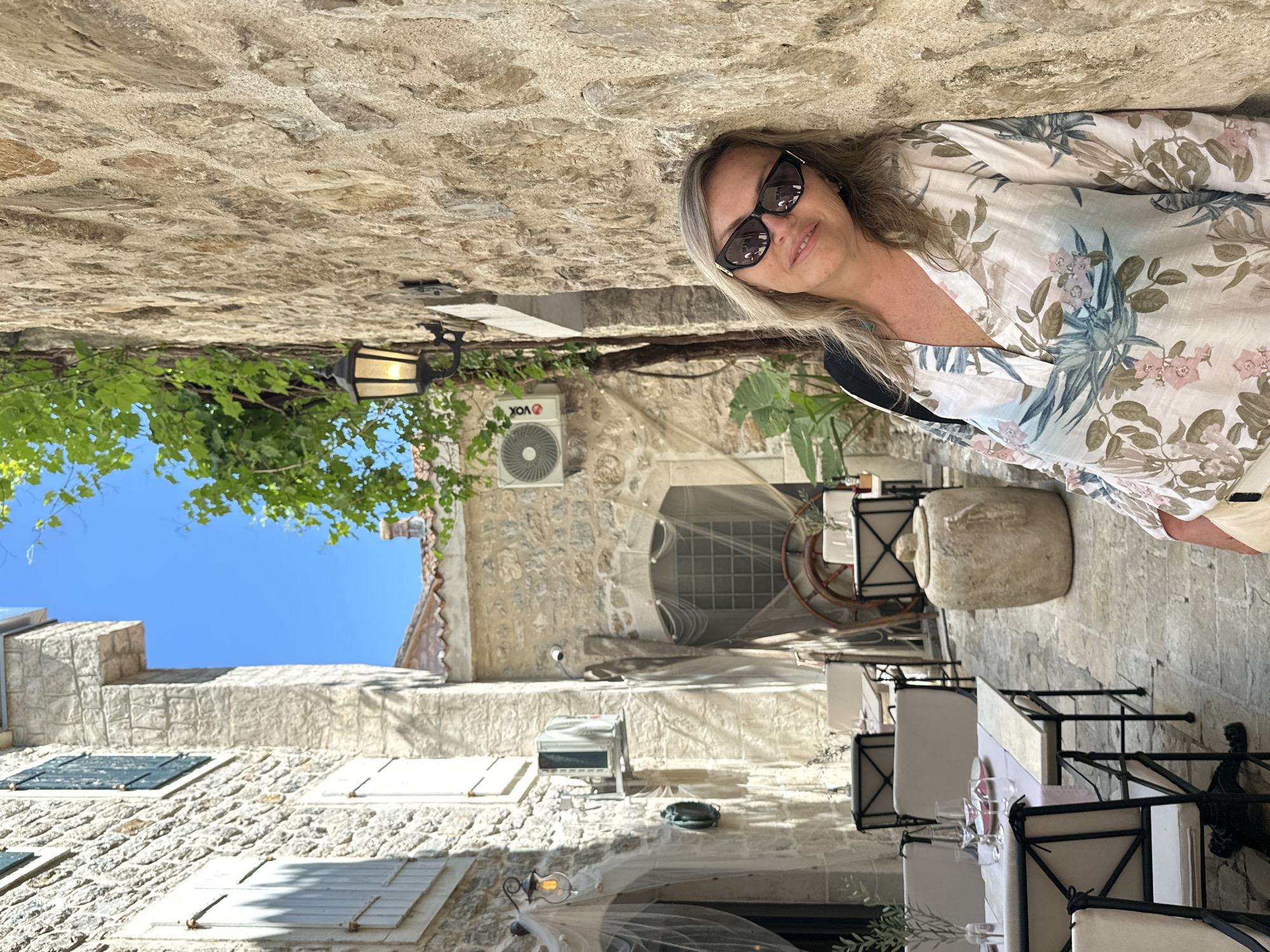 A medium shot captures a woman in sunglasses, leaning against a stone wall in the city of Budva, Montenegro. She's wearing a floral blouse and standing in a narrow street, where tables are set for dining. Stone buildings line the street, and an air conditioning unit is visible on one of them. A street lamp and some foliage hang overhead. The sky is visible in the distance.