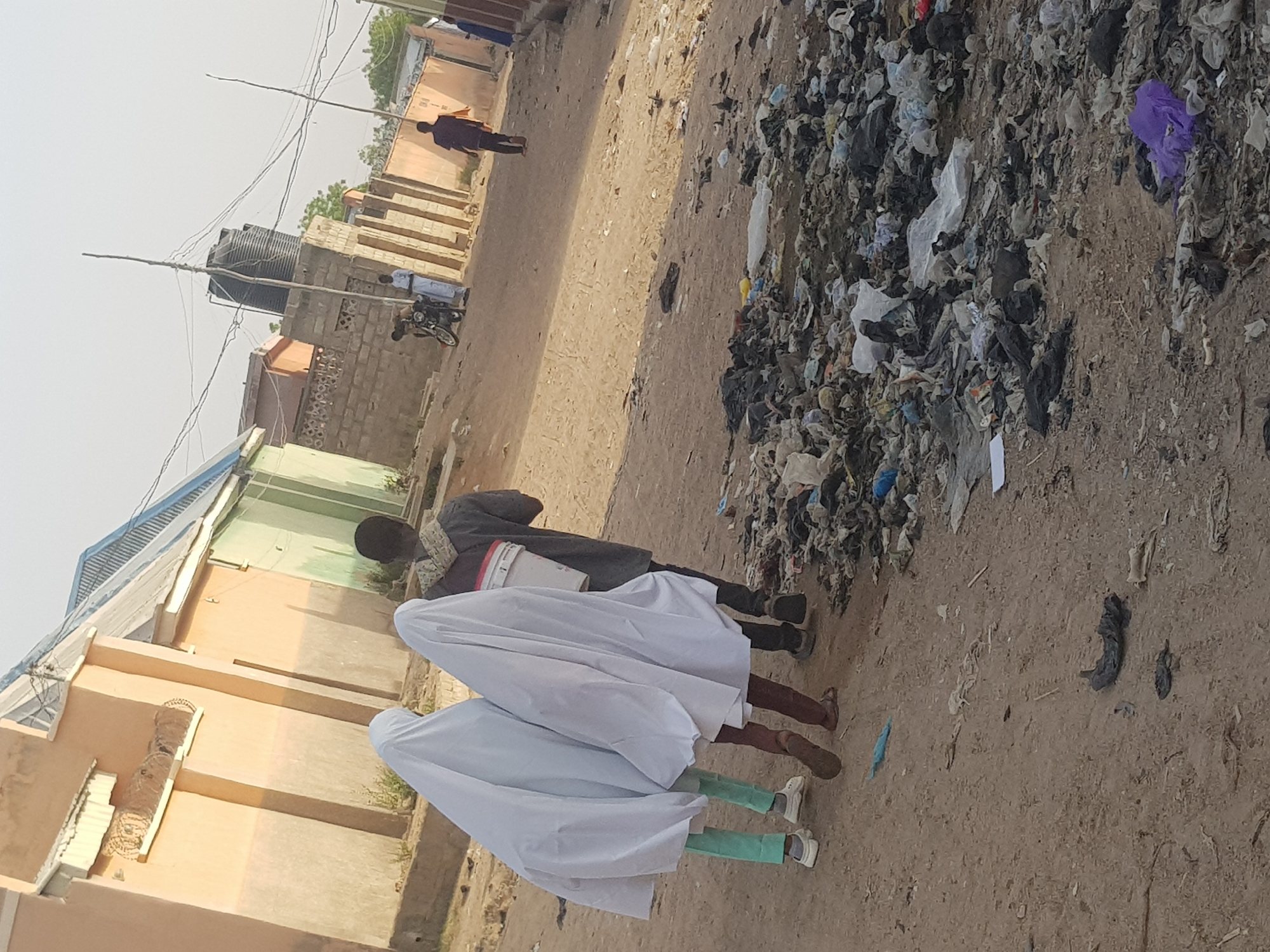 The image shows a street in Dutsinma B, Nigeria. Two people wearing white garments that cover their heads and bodies walk alongside a person wearing casual clothes. A pile of trash is next to them. In the background are buildings and a person walking along the street.