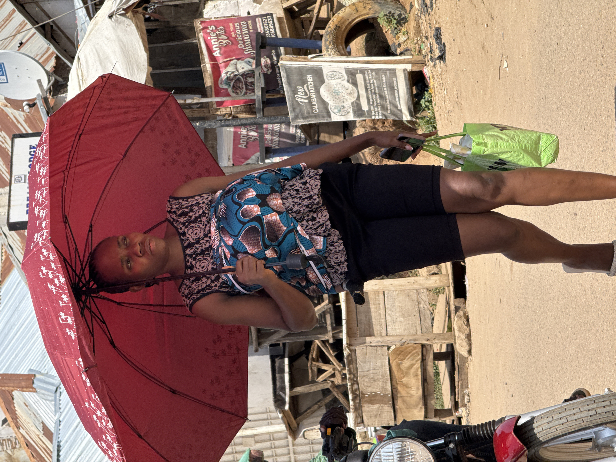 A woman walks under a red umbrella in Abuja, Nigeria, holding a green tote bag in her right hand and a black mobile phone in her left hand. She is wearing a sleeveless printed top and black shorts. The background features market stalls, corrugated iron roofs, a satellite dish, a motorcycle, wooden crates, and a discarded tire. Signs for "Annie's Bites Delicious Shawarma" and "New Calabar Kitchen" are visible. The ground is sandy and dusty.