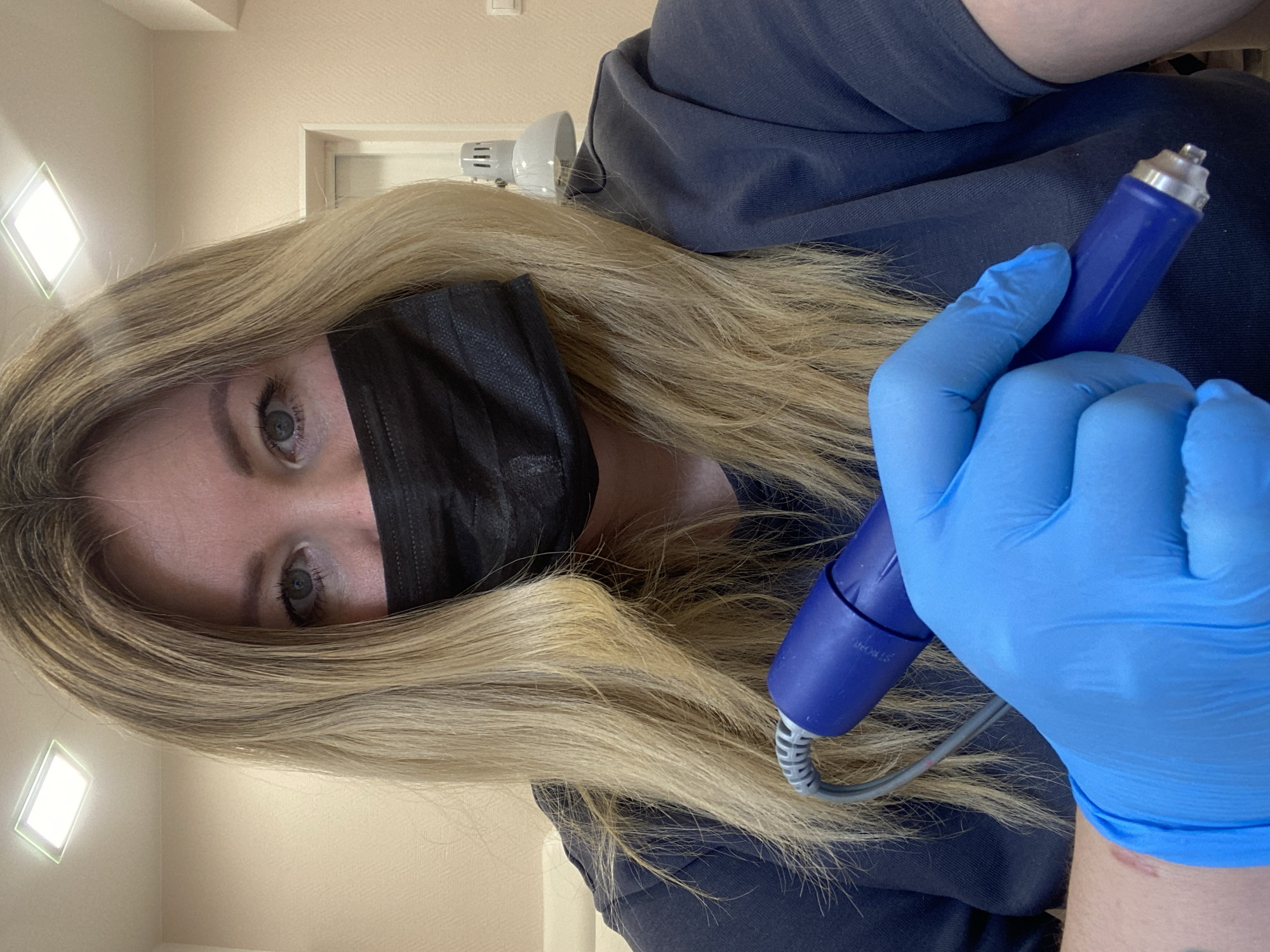 A woman wearing a black face mask and blue nitrile gloves is holding a blue rotary tool. Her long, blonde hair is partially covering her face. She is wearing a navy blue shirt. The background includes a wall with a square light fixture, a wall-mounted lamp, and a door frame. There is no verifiable information linking this image to the city of Chaikovsky, Russia.