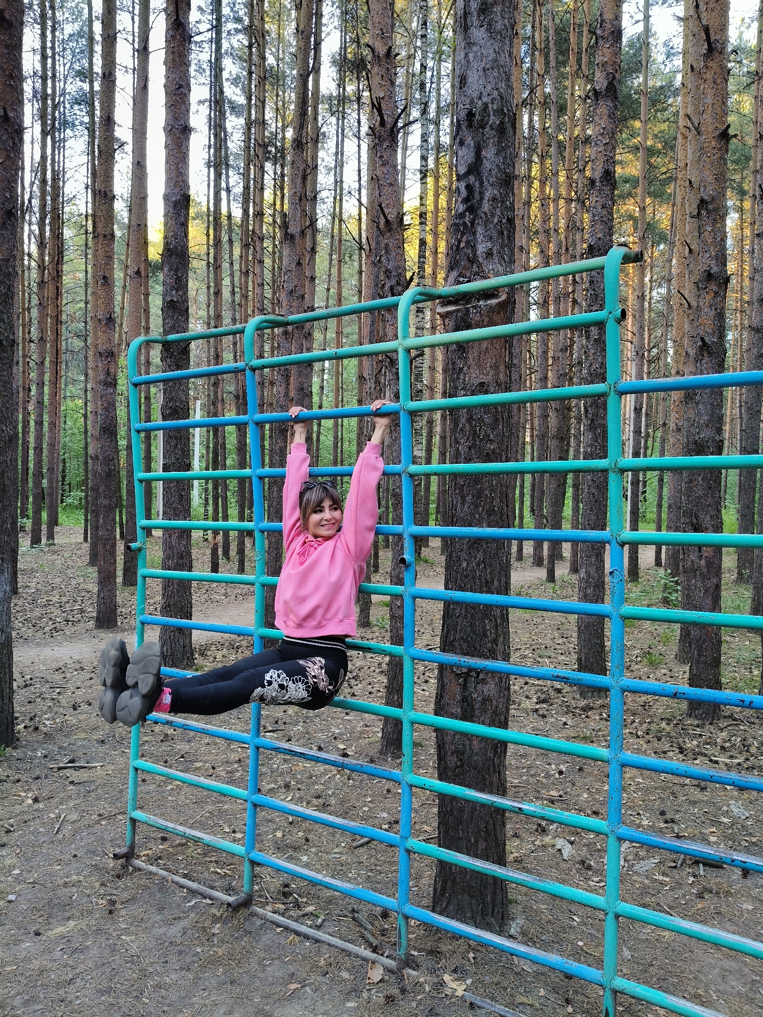 A girl is hanging on a blue and green metal climbing frame in a forest in Chaikovsky, Russia. She is wearing a pink hoodie, black leggings with white flower designs, and gray sneakers with pink socks. The climbing frame is situated among tall pine trees. The ground is covered with dirt, pine needles, and pine cones.