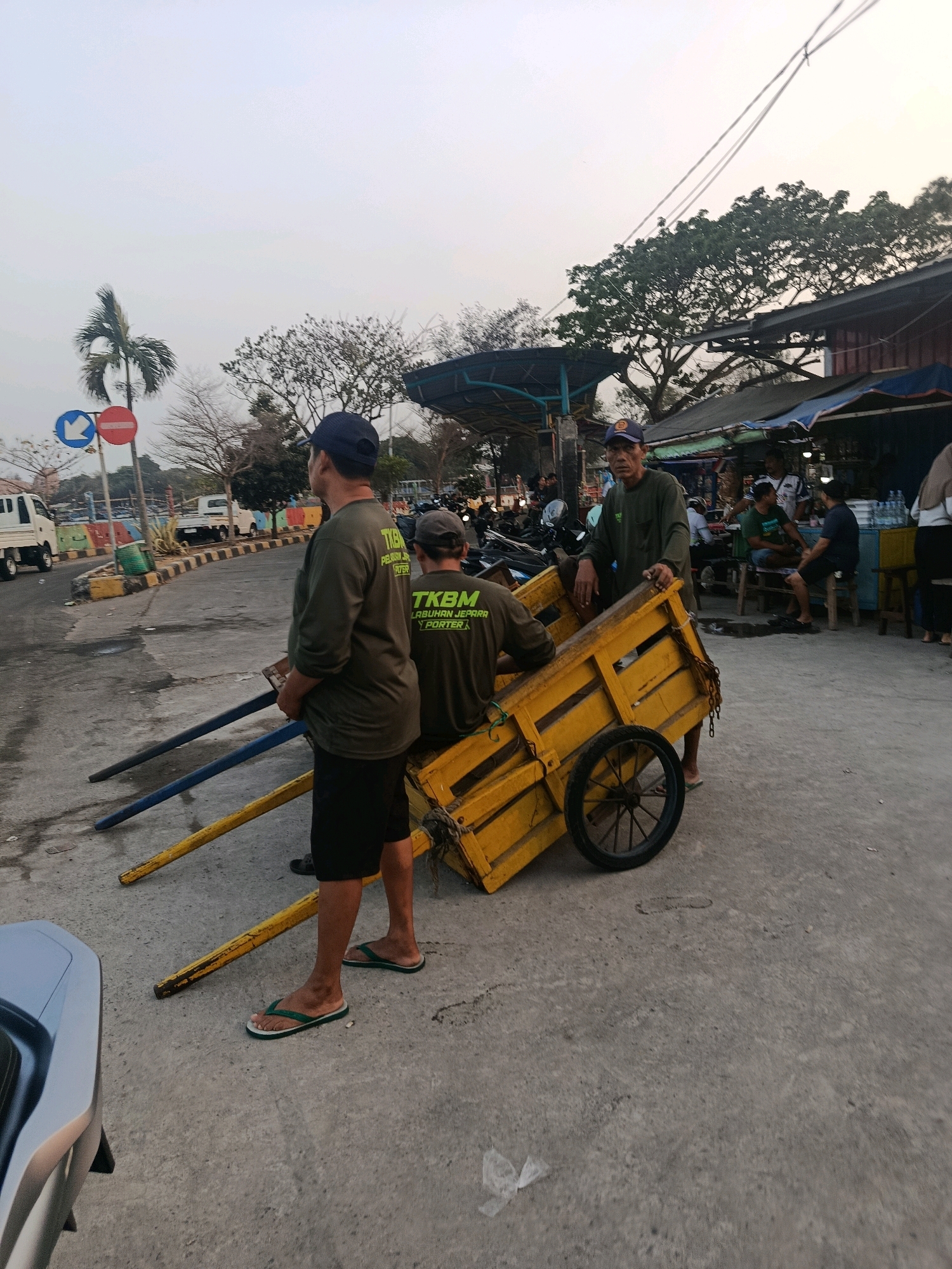 The image depicts a street scene in Jepara, Indonesia, likely near a port or market area, as suggested by the presence of a yellow cargo cart, people in work attire, and what appear to be food stalls in the background. Three men are visible. One man in the foreground, wearing a dark blue cap and a green long-sleeved shirt with "TKBM PELABUHAN JEPARA PORTER" printed on it, stands facing away from the camera, looking towards the right. He is wearing black shorts and green flip-flops. To his right, another man, also in a green long-sleeved shirt with the same text, is seated on the yellow cart, also facing away. He wears a dark grey cap. A third man, wearing a green long-sleeved shirt and a blue baseball cap, is standing behind the cart, facing the camera and looking slightly to the left. He is operating the cart.
The yellow cart is wooden with metal reinforcement and has a single spoked wheel. Two blue poles are positioned in front of it, possibly for stabilization or as part of a loading mechanism. In the background, there are several stalls with awnings, motorbikes, and other people. Traffic signs, including a "no entry" sign and a blue arrow sign, are visible on the left side of the road. The sky is overcast, suggesting early morning or late afternoon. The overall environment appears to be a bustling area associated with daily commerce and labor.