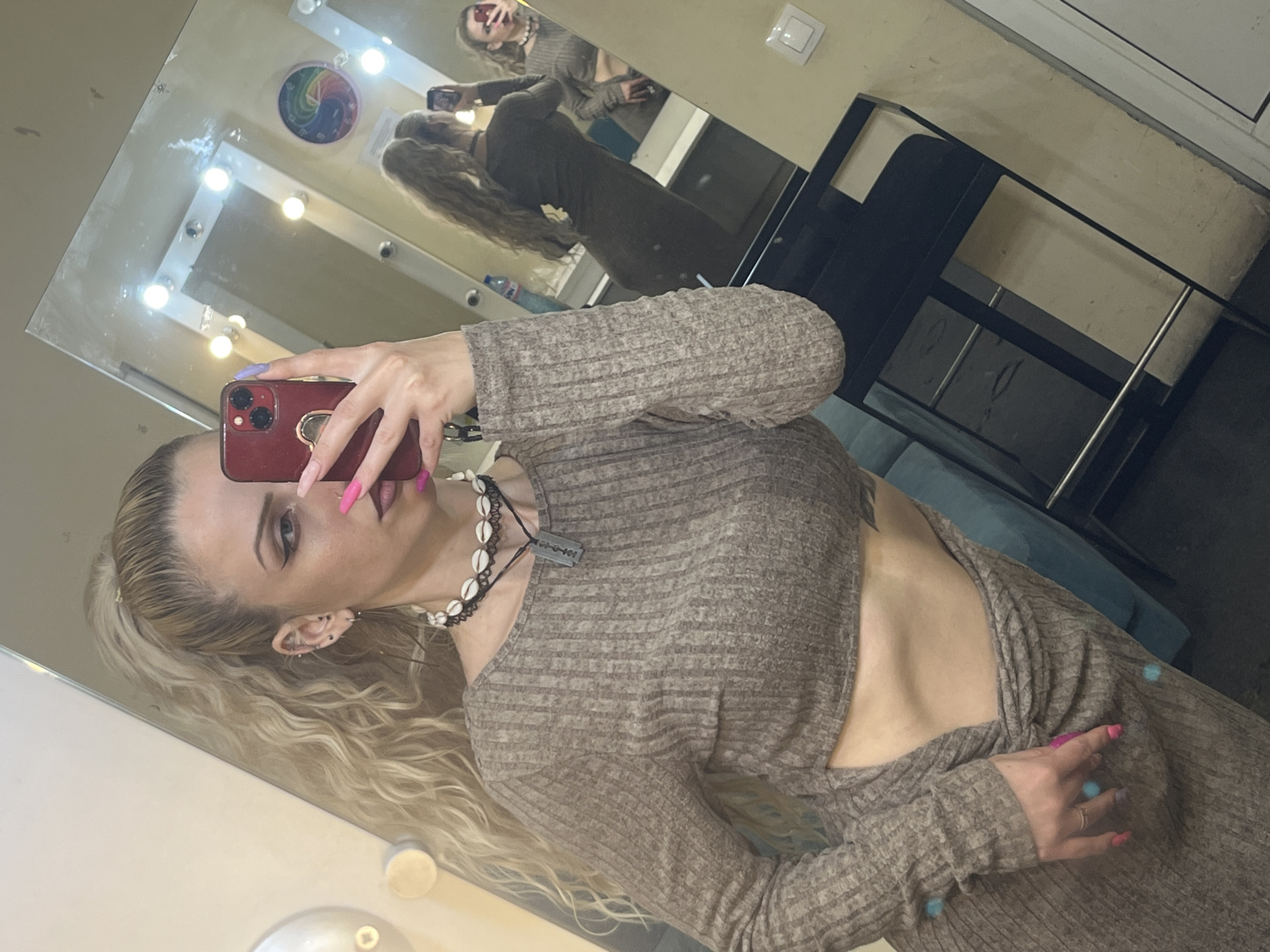 A person is taking a selfie in a room. The person has long, blonde, curly hair pulled back in a ponytail, fair skin, and several piercings in their left ear. They are wearing a brown long-sleeved dress with a cutout at the waist, revealing their bare midriff. A necklace with seashells and a metallic pendant adorns their neck. The person is holding a red smartphone with multiple cameras, and their fingernails are painted with various shades of pink and purple.
In the background, a vanity mirror with lightbulbs is visible. The reflection in the mirror shows the person taking the selfie, as well as a glimpse of a chair and a section of the room. A colorful, circular decoration is hanging on the wall behind the mirror.
The location cannot be determined to be in Plovdiv, Bulgaria, based on the information present in the image.