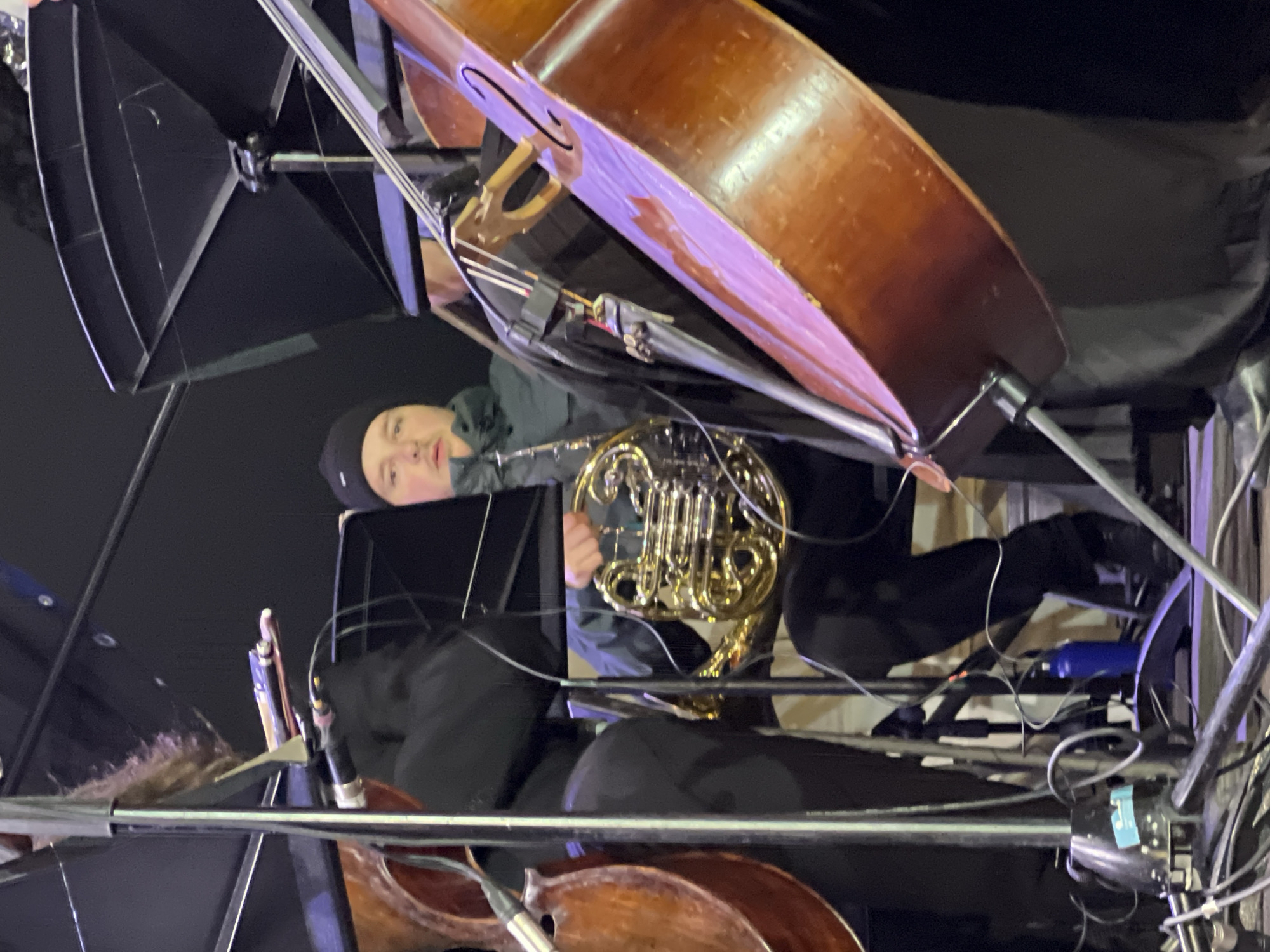 A person in a black beanie and dark clothing holds a French horn amid a complex arrangement of musical instruments, stands, and wires. There is a large double bass, and the scene includes music stands and cables. The location is undetermined, and there is no visible evidence to indicate that it is in the city of Saint Petersburg, Russia.