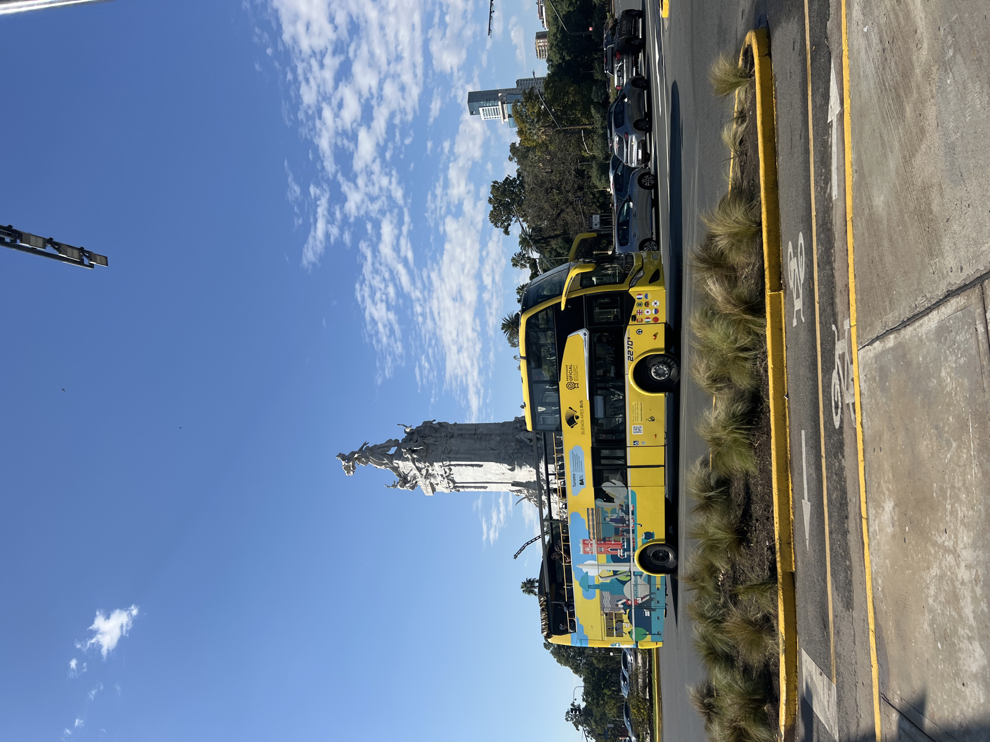 A yellow double-decker tourist bus with the words "Buenos Aires BUS" is parked on the side of a road next to a bike lane, with the Monumento de los Españoles in the background. The sky is blue with scattered clouds. Cars are parked further down the road, and some buildings can be seen in the distance. The bus has various images on its sides, including flags and cityscapes. The number 2270 is on the bus.