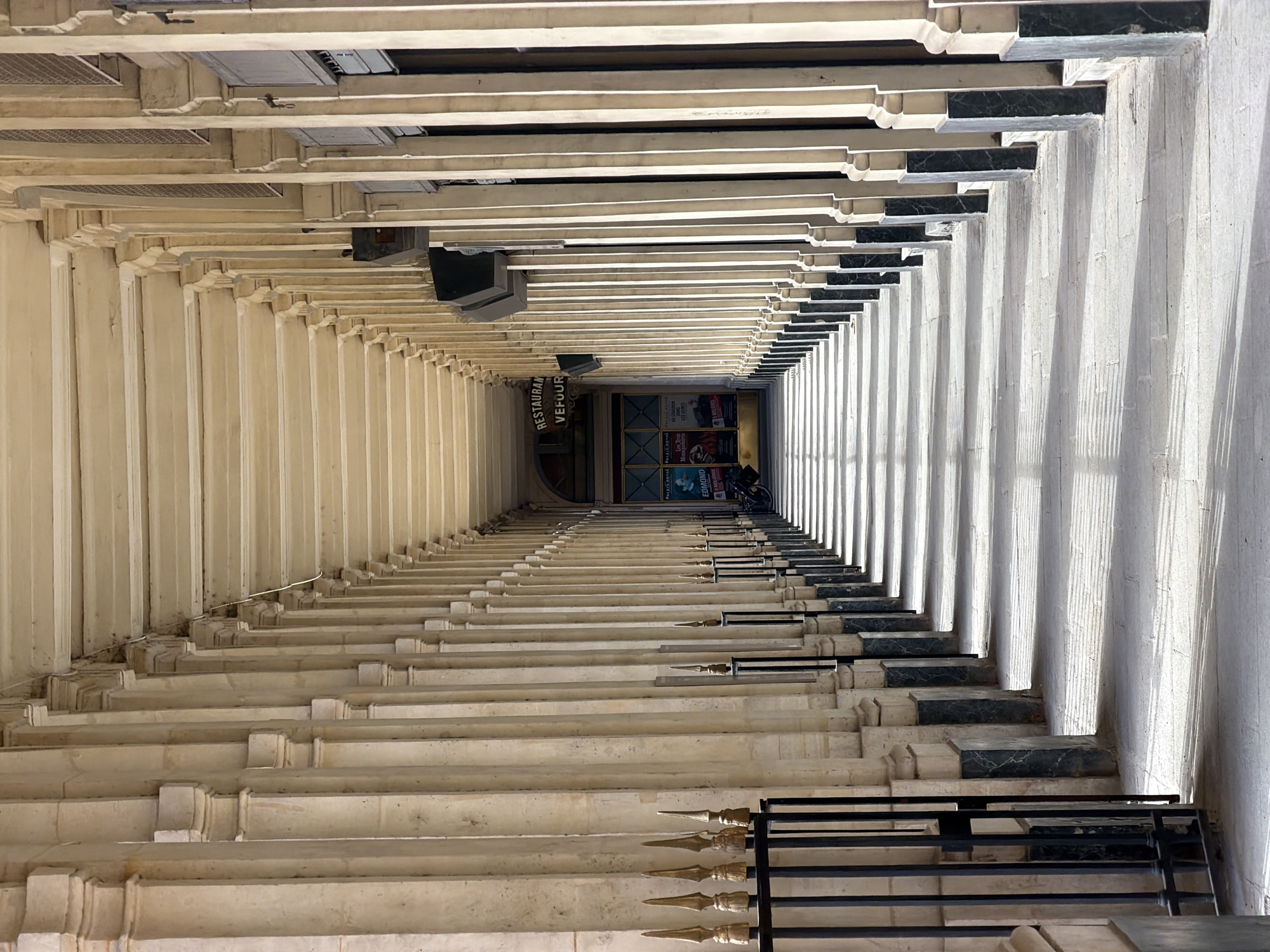 The image presents a striking high-angle perspective looking down a long, grand arcade or gallery, likely part of the Palais-Royal in Paris, France. The scene is dominated by repeating architectural elements: numerous light-colored, ornate pillars and moldings that create strong converging lines, emphasizing depth. On the right side, dark, polished marble-like blocks cap some sections of the structure, providing a visual contrast to the predominant beige stone. In the immediate foreground, a dark metal railing adorned with golden spiked finials is partially visible.
Strong, directional sunlight from the right casts dramatic, elongated vertical shadows from the columns across the passage, highlighting the texture of the stone and indicating a clear, bright day. At the far end of the arcade, a building facade comes into view, displaying various posters and a prominent sign. Two indistinct figures can be seen near the entrance at the distant end.
Visible text in the image includes "PALAIS ROYAL" (appearing twice, once as "PALAIS ROTAL" in OCR), and "RESTAURAN VEFOUR" (referring to the Grand Véfour restaurant). Other text snippets from posters include "Les T...", "Mosquelaires", "EDMOND", "DO CHARBON", and "LES VEINES". The overall impression is one of elegant, classical architecture and profound perspective.