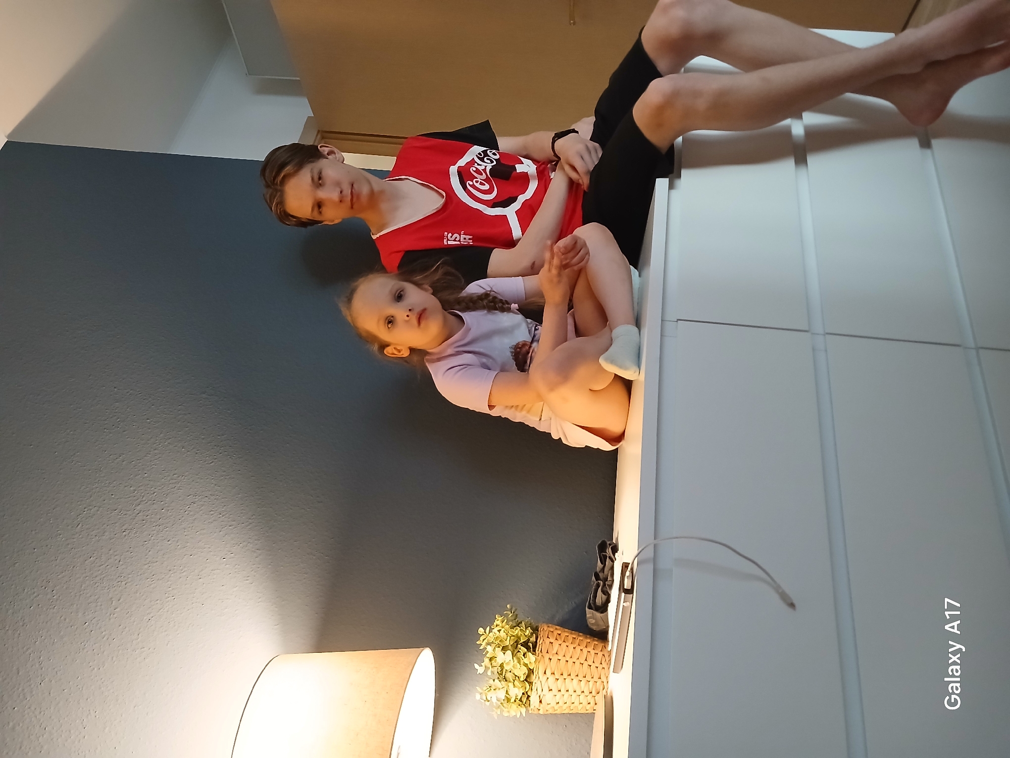 Two children are sitting on a white chest of drawers in a room. The older child, a teenage boy, is wearing a red Coca-Cola t-shirt and black shorts. The younger child, a girl, is wearing a lavender t-shirt and is barefoot. A table lamp with a lampshade and a decorative plant in a wicker basket are visible on top of the chest of drawers. The wall is painted in a dark gray color. The image includes "Galaxy A17" watermark. The location is not discernible from the provided image.