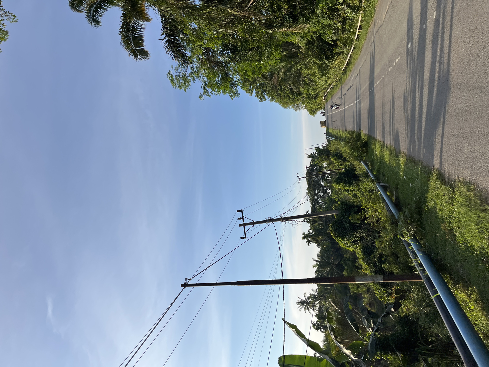 This is a photograph taken in Sukaramai Dua, Indonesia, depicting a paved road ascending a gentle incline. The sky is a clear, bright blue with a few wispy clouds. The road is bordered on the left by dense green foliage and several utility poles with wires, and on the right by a lush green embankment with trees and palm trees. There are two motorcyclists visible on the road, one further ahead than the other, both traveling uphill. A truck is also visible in the distance at the crest of the hill. The lighting suggests it is daytime, possibly late afternoon, with long shadows cast across the road. The overall impression is one of a rural or semi-rural road in a tropical environment.
