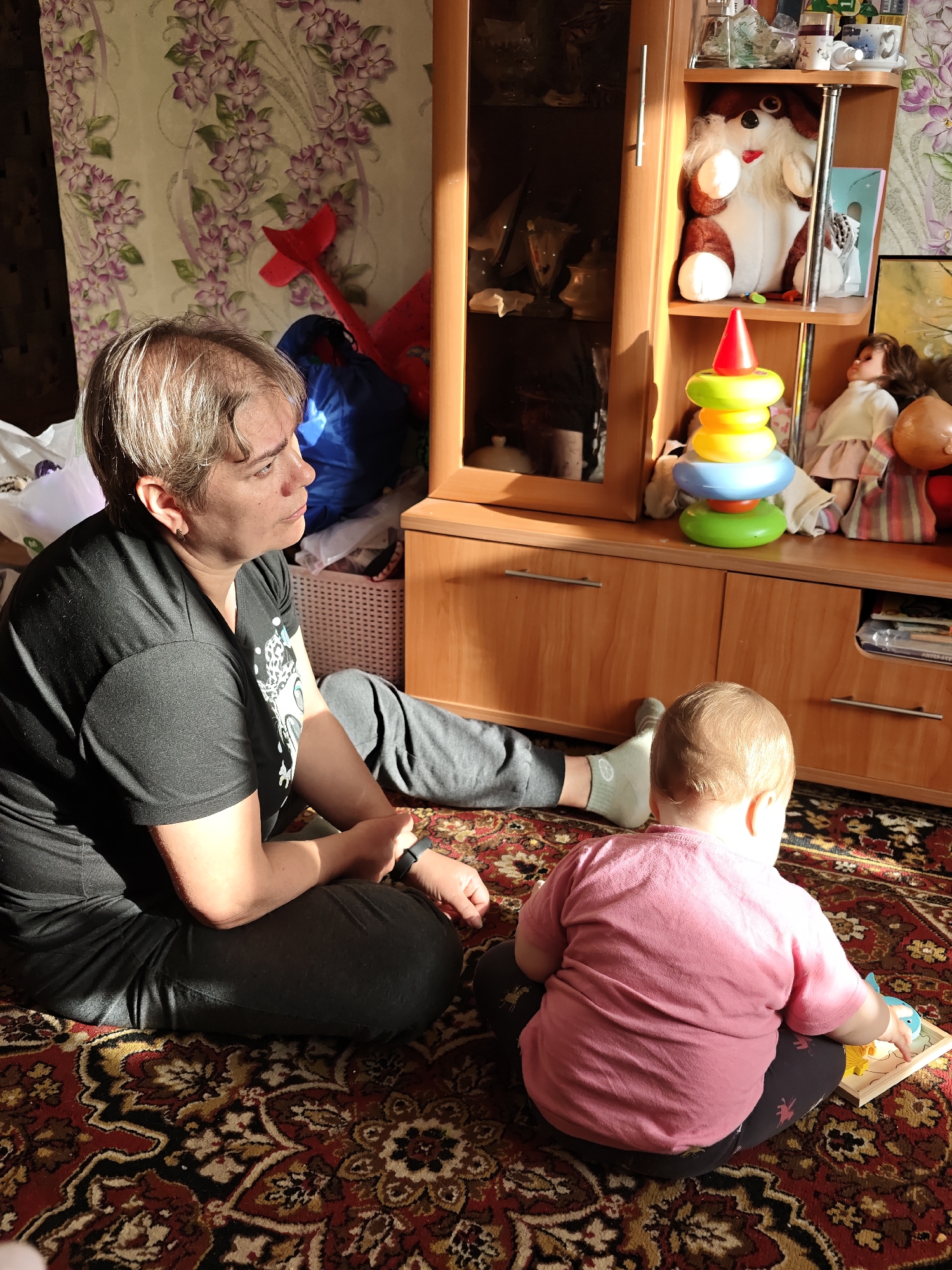The image captures a domestic scene in Уральское, Russia. A middle-aged woman with short, graying hair, wearing a black t-shirt and dark pants, sits cross-legged on a patterned rug, looking towards a young child. The child, facing away from the camera, is wearing a pink shirt and dark pants and is engaged with a toy or puzzle on the rug. Behind them is a wooden cabinet containing toys, including a stuffed dog, a colorful ring stacker, and a doll. To the left of the cabinet, wallpaper with a floral pattern is visible, along with some bags and a red toy. The scene is illuminated by sunlight entering the room.