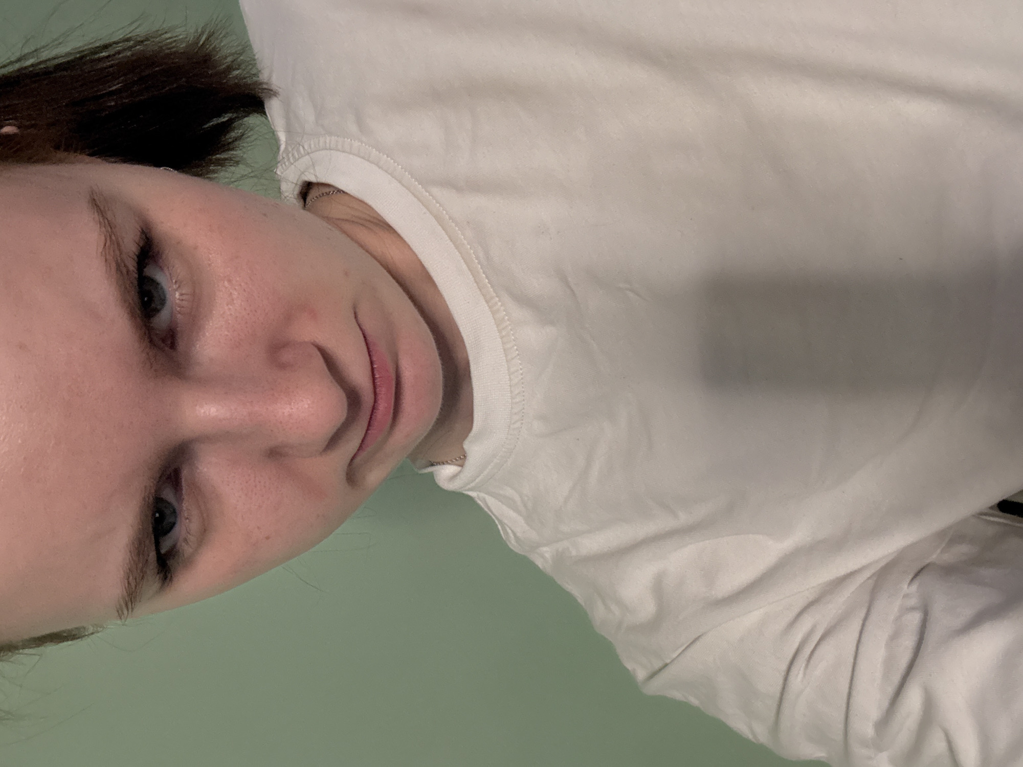 A close-up, tilted portrait shows a person with fair skin, brown hair, and blue eyes. The background is a solid light green color. The person is wearing a white t-shirt with a crew neck. A thin chain necklace is visible around the person's neck. The location is Unknown, Russia.