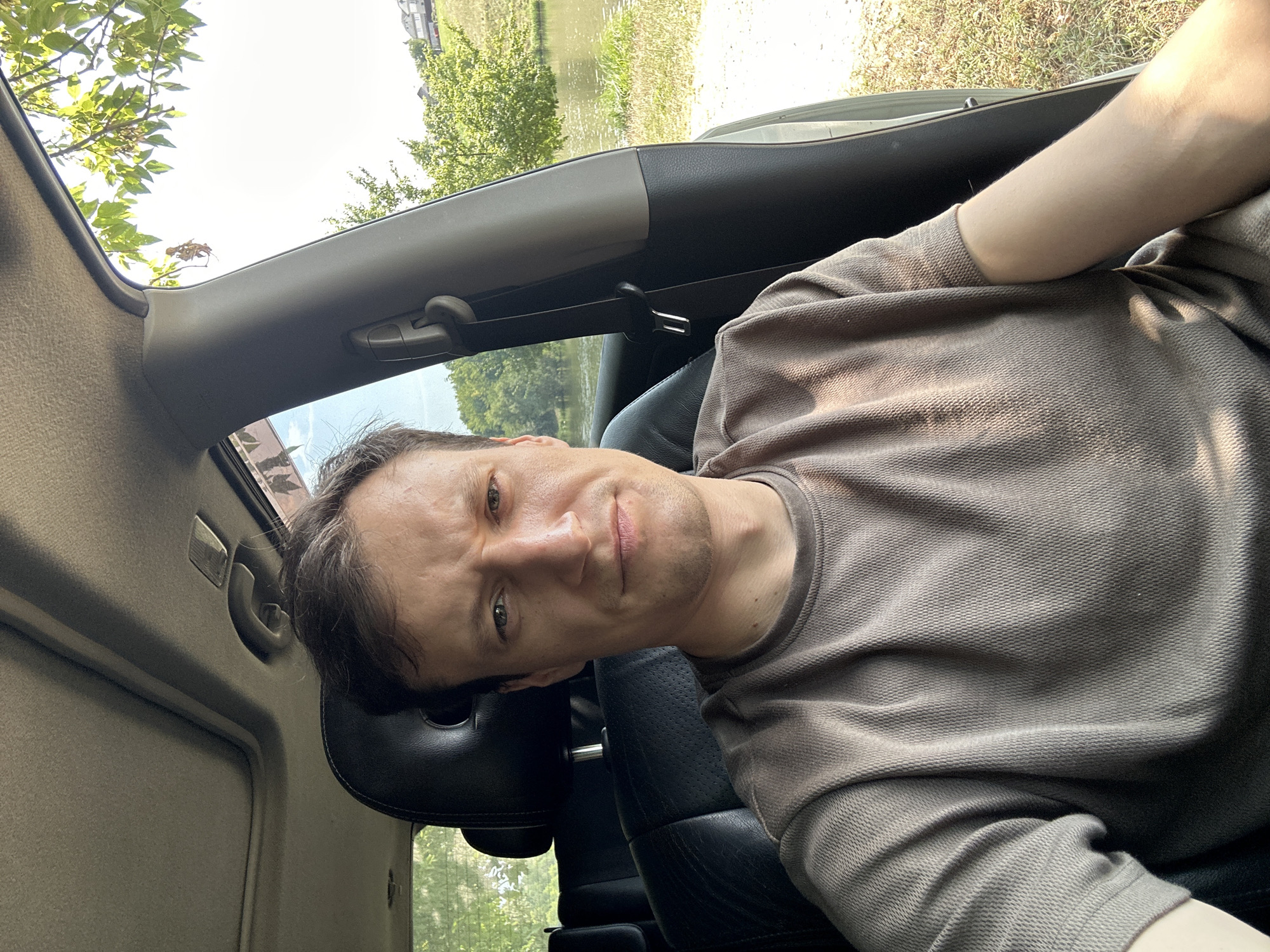 A man is sitting in the driver's seat of a car. He is looking directly at the camera with a slightly furrowed brow. He has short, dark brown hair and is wearing a light brown t-shirt. The interior of the car is visible, including the black leather seat and the car's ceiling. Through the open car window, a body of water, trees, and houses on a hill are visible. The scene appears to be a sunny day. The location is identified as Makiv, Ukraine.