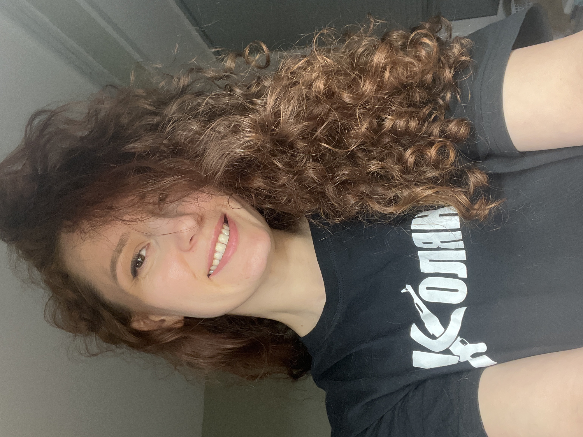 A woman with curly, light brown hair is smiling at the camera. She is wearing a black t-shirt with a white graphic print that includes the word "KOUTANA". The background is a neutral-toned wall. The location is not evident from the image.