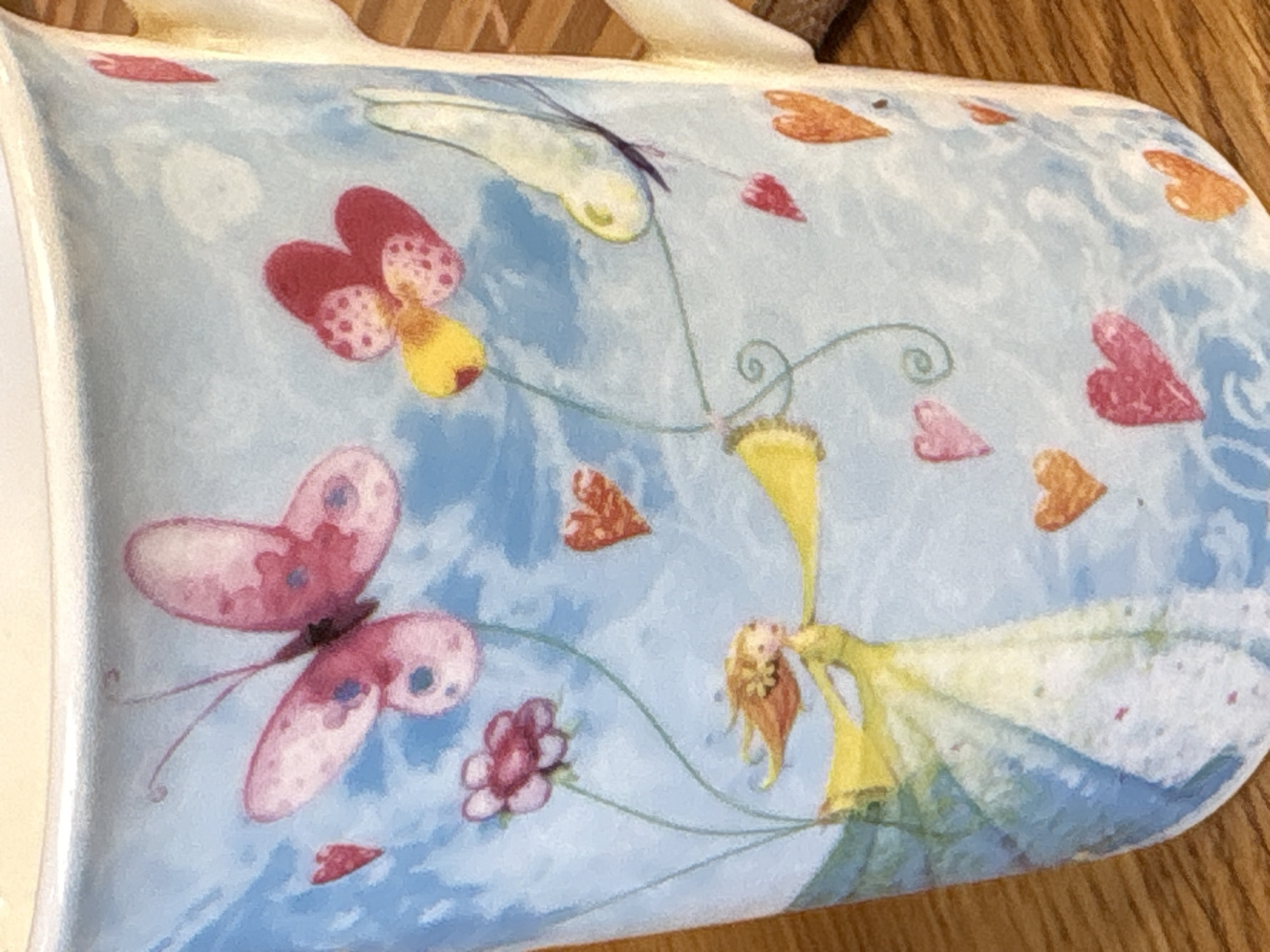 The image depicts a portion of a light blue mug with a painted design. The design features butterflies with pink and purple markings, red and orange hearts, a pink flower, and a fairy with blonde hair wearing a yellow and white dress. The fairy appears to be hanging from a yellow bell-shaped flower. The mug has a white rim and handle. The background is a wooden surface. There is no indication of the location being Ленинградская, Russia in the image.