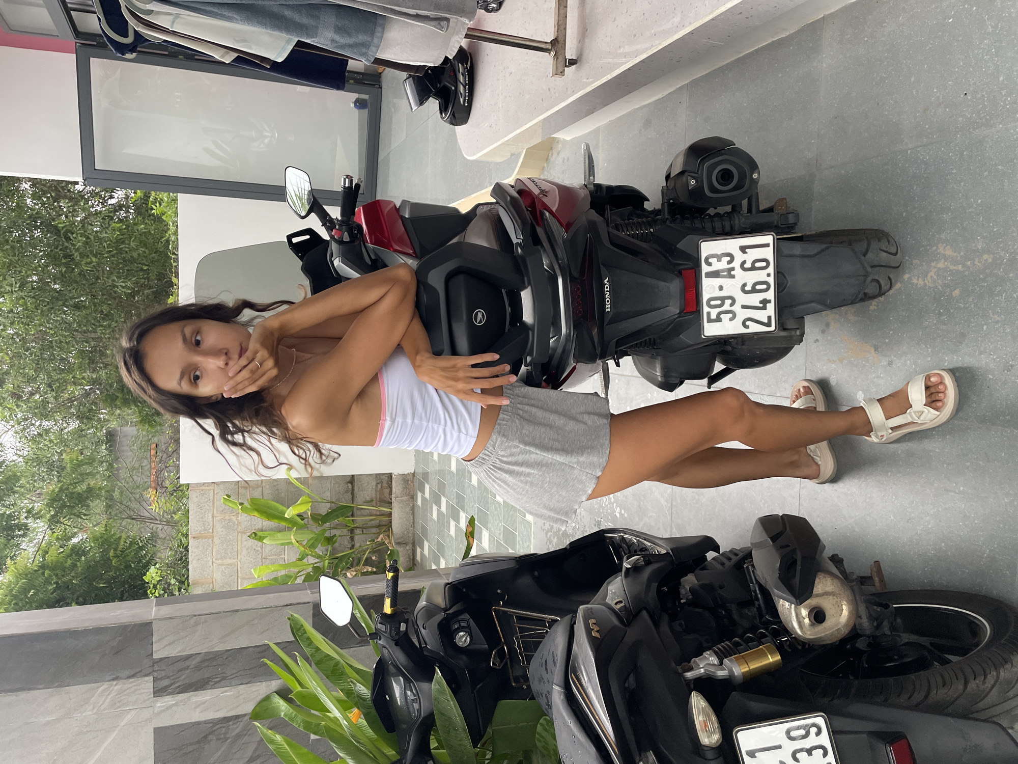 A young woman is standing next to a red scooter in Mũi Né, Vietnam. She is wearing a white crop top with pink trim and a gray skirt. She has her right arm resting on the back of the scooter and her hand on her chin, looking away from the camera. Another black scooter is parked to her left. The background shows a tiled wall, some greenery, and a clothing rack. The floor is tiled with a gray and white pattern. The license plate on the red scooter reads "59-A3 246.61".