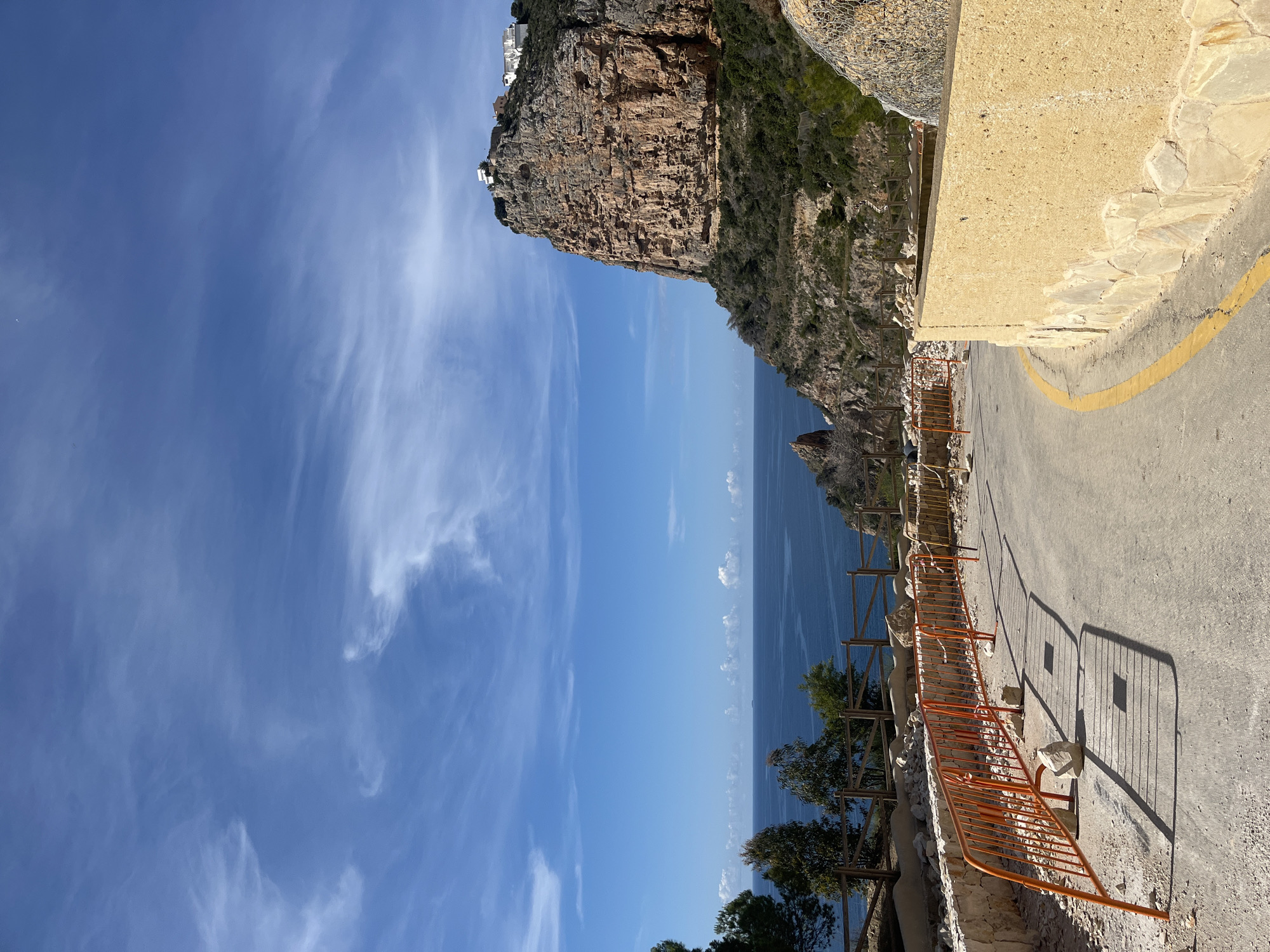 The image captures a scenic view in El Poble Nou de Benitatxell / Benitachell, Spain. A rugged cliff face, topped with some buildings, dominates the left side of the frame. Below the cliff, the blue sea stretches towards the horizon. An elevated pathway runs along the right side, with a yellow safety line painted near the edge. An orange metal railing is visible along the pathway, casting a shadow on the paved surface. Trees and vegetation grow along the cliffside below the pathway. The sky is a clear blue with whispy clouds.
