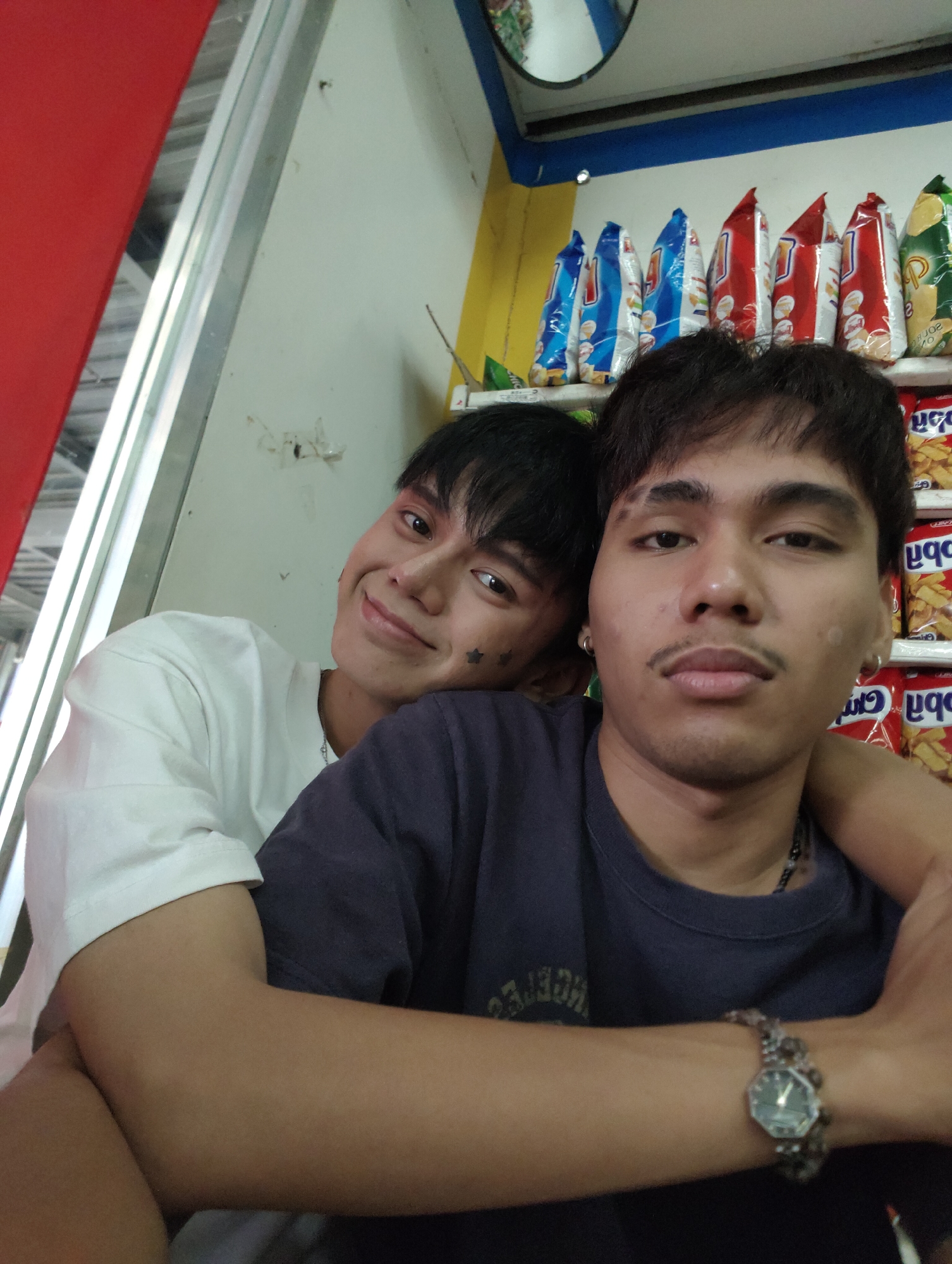 The image shows two young men posing for a selfie. The man in the foreground on the right, wearing a dark shirt and a watch, has his arm around the man on the left, who is wearing a white shirt and has a star-shaped tattoo on his cheek. They are in what appears to be a small convenience store, with shelves of snacks visible in the background. The store's interior is brightly lit, with red and yellow accents on the walls and a blue stripe along the top. A mirror is visible on the ceiling. The overall impression is of a casual, candid moment captured in a local shop.