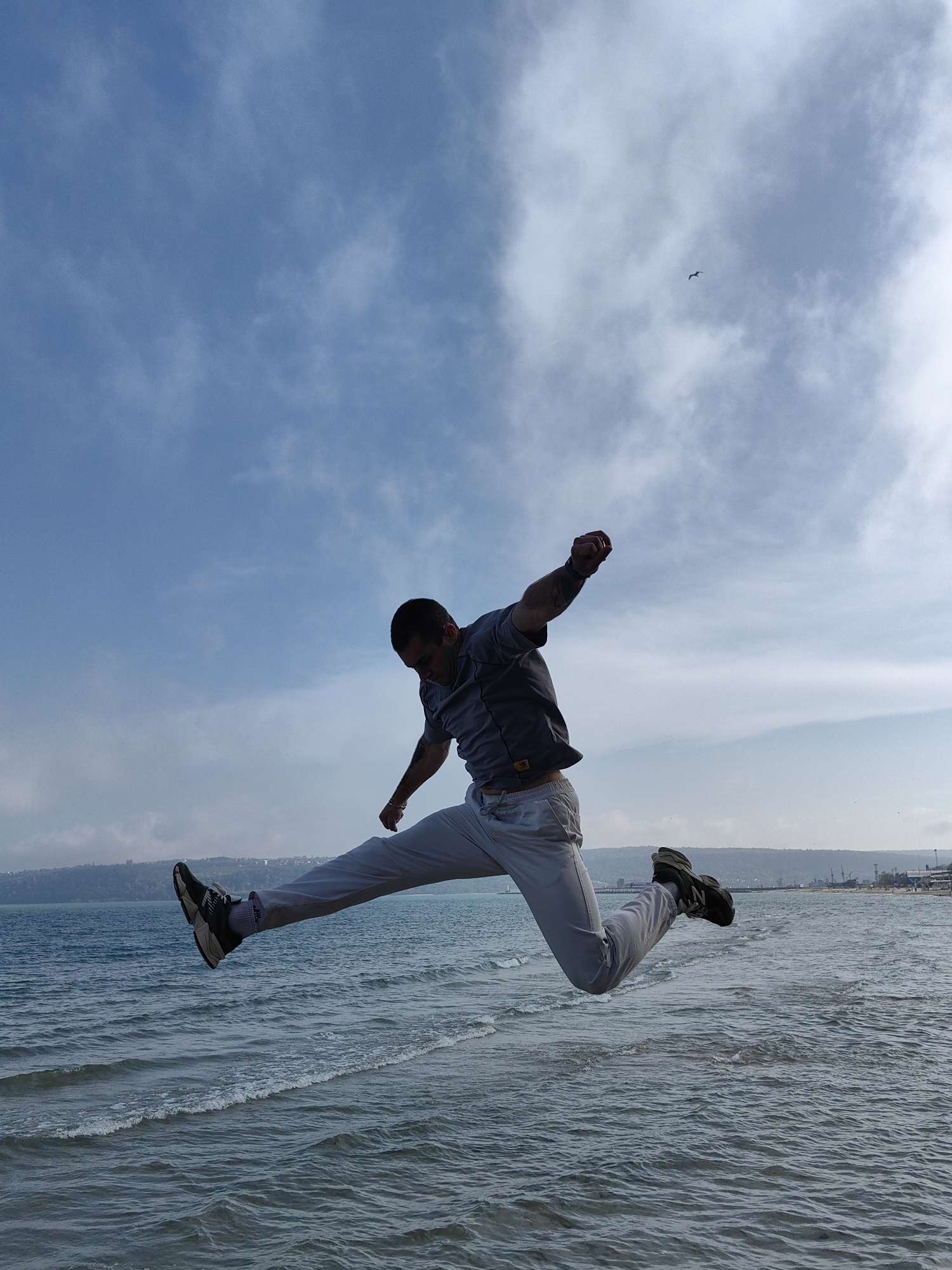 A man jumps in the air in front of the sea in Varna, Bulgaria. The man is wearing a grey t-shirt, light-colored pants, and athletic shoes. His right arm is raised in a fist. The sea is in the background with a coastline and buildings visible in the distance. The sky is blue with scattered clouds, and a bird is flying in the upper part of the frame.
